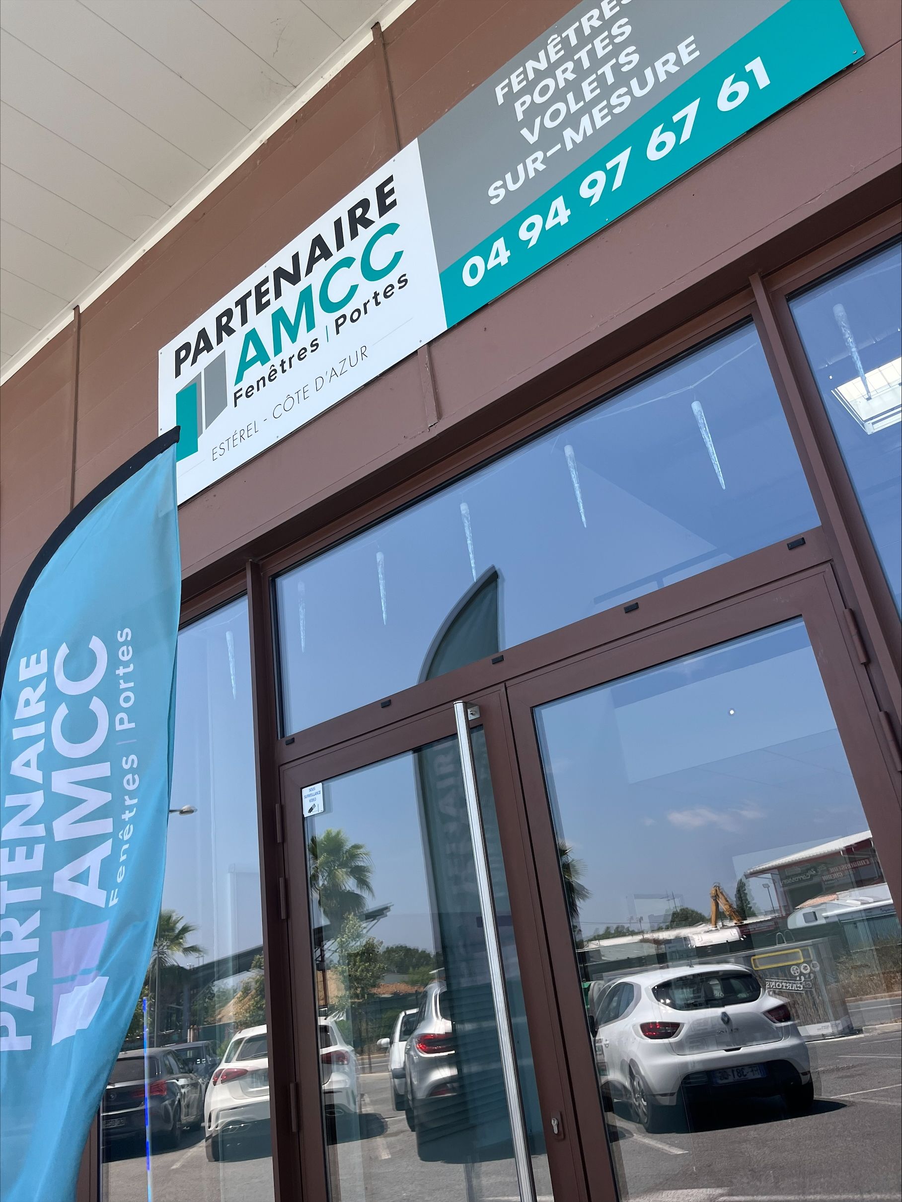 Enseigne d'AMCC, entreprise de fenêtres et de portes, au-dessus d'une vitrine. Un drapeau publicitaire bleu est hissé devant.