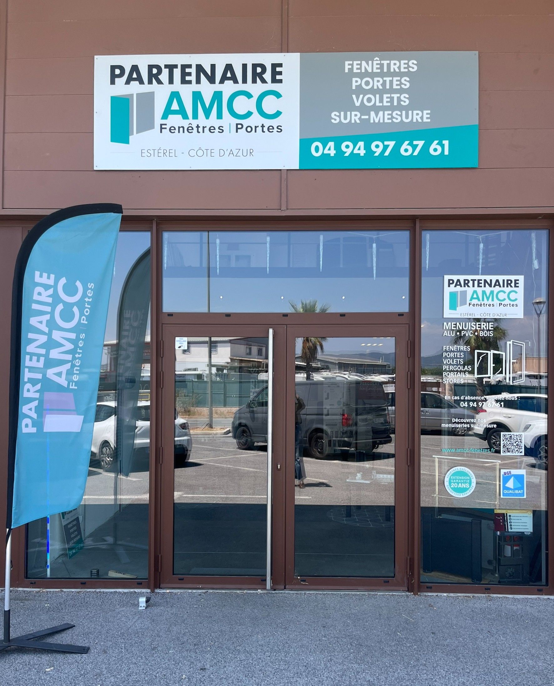 Enseigne d'une entreprise de fenêtres et de portes (AMCC), avec le numéro de téléphone 04 94 97 67 61, au-dessus de l'entrée vitrée. Un drapeau turquoise est visible à gauche.