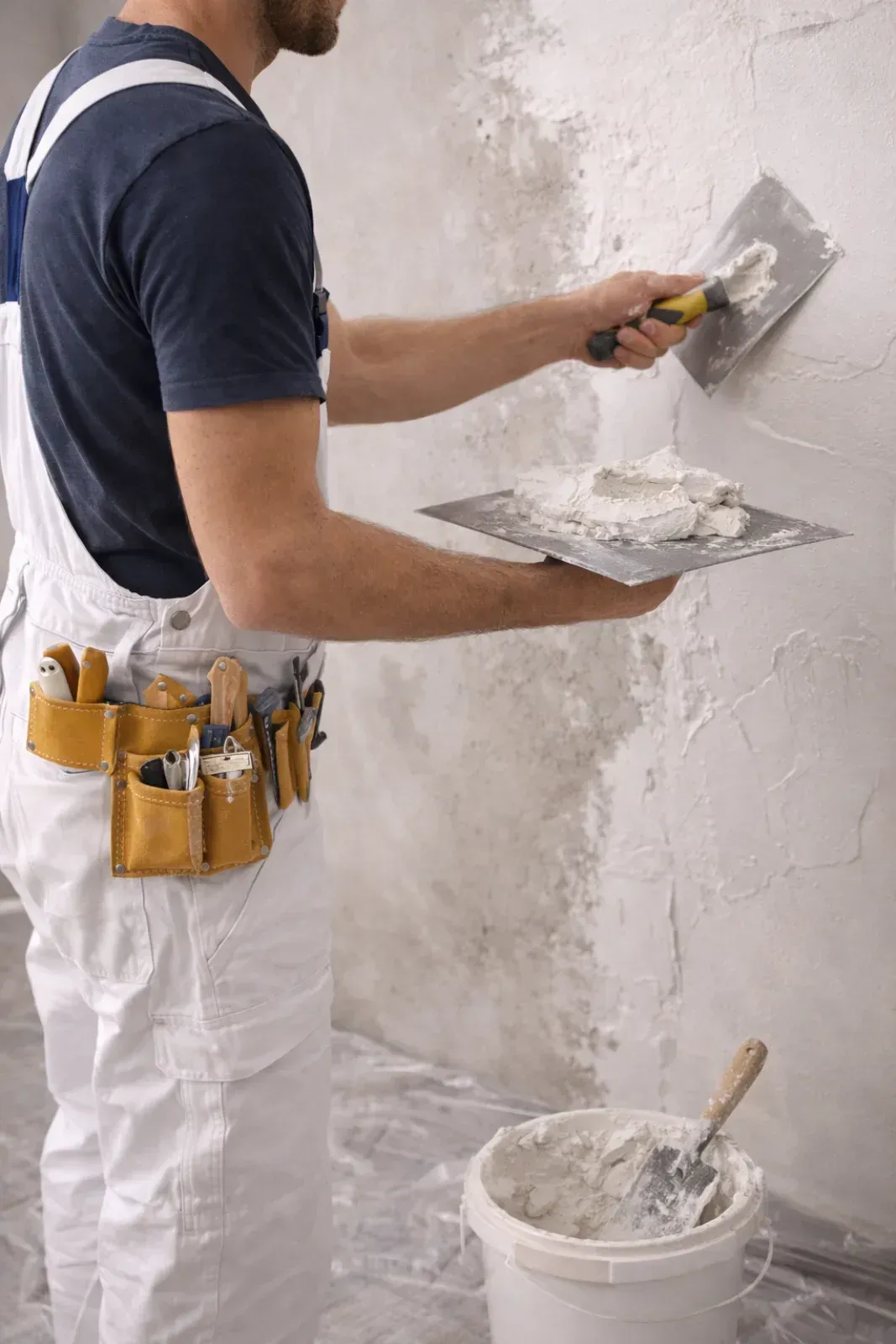 Eine Person trägt mit einer Kelle Gips an einer Wand auf und hält eine Gipswanne. Sie trägt einen weißen Overall.