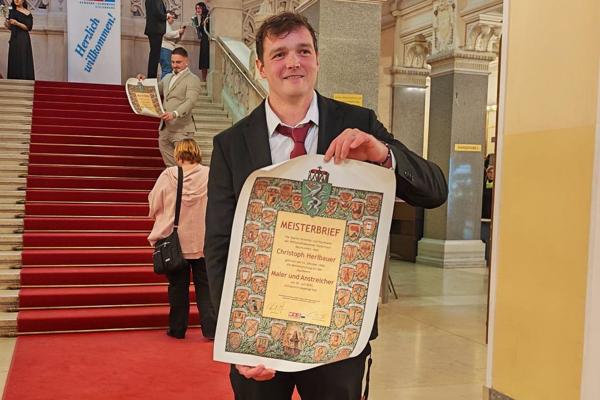 Ein Mann im Anzug hält ein verziertes Dokument in der Hand und steht auf einer mit rotem Teppich ausgelegten Treppe in einem prunkvollen Gebäude.