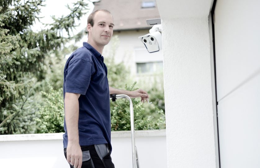 Mann im blauen Hemd in der Nähe einer Überwachungskamera; hält eine Leiter; vor einem weißen Gebäude mit Grünflächen.