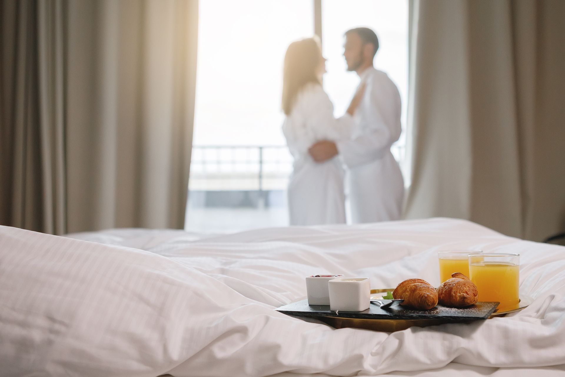 Plateau de petit-déjeuner sur le lit d'une chambre d'hôtel, avec un couple en peignoirs blancs debout près de la fenêtre en arrière-plan.
