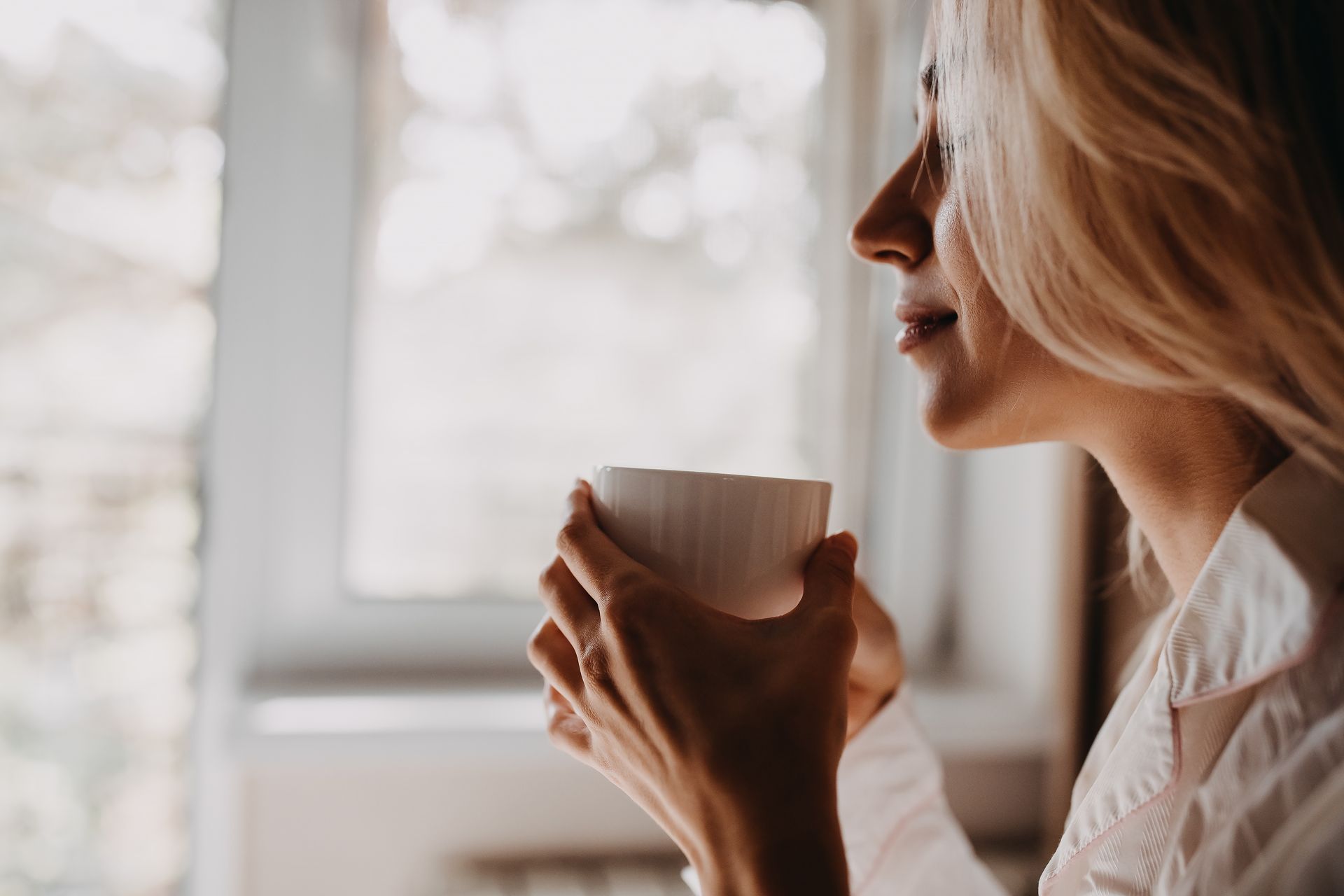 Une personne en chemise blanche tenant une tasse près d'une fenêtre, regardant au loin dans la douce lumière du jour.