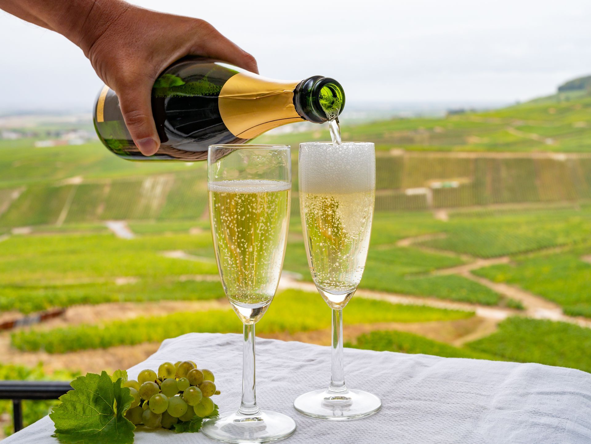 Versement manuel de vin mousseux dans deux flûtes à champagne, avec des terrasses de vignes en arrière-plan.