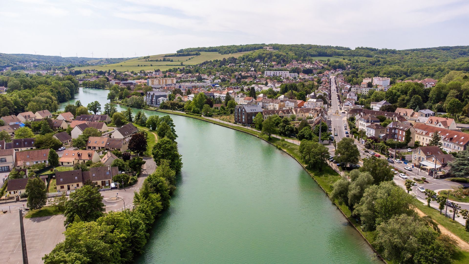 Vue aérienne d'une rivière serpentant à travers une ville, avec des maisons, des arbres et des collines en arrière-plan.