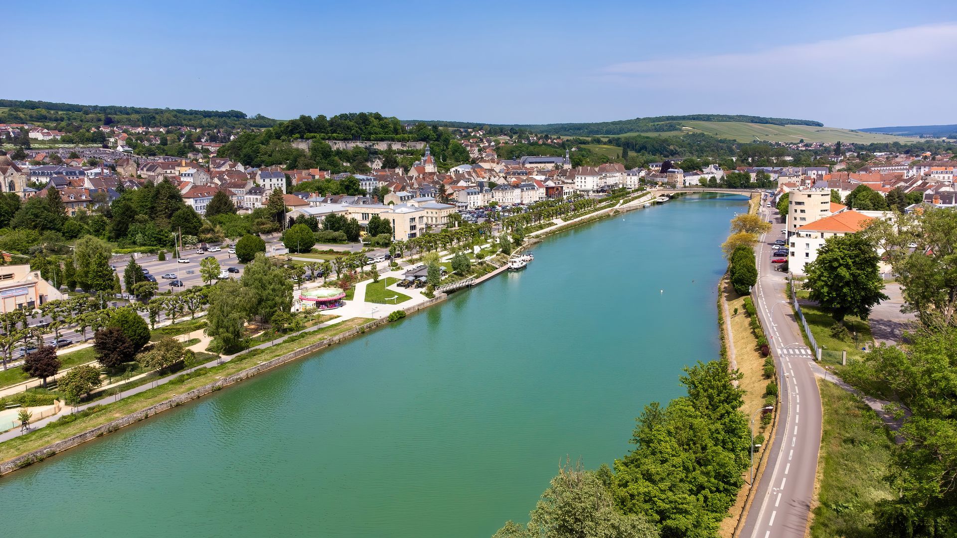 Vue aérienne d'une rivière verdoyante traversant une ville avec des routes, des maisons et des arbres le long des berges.