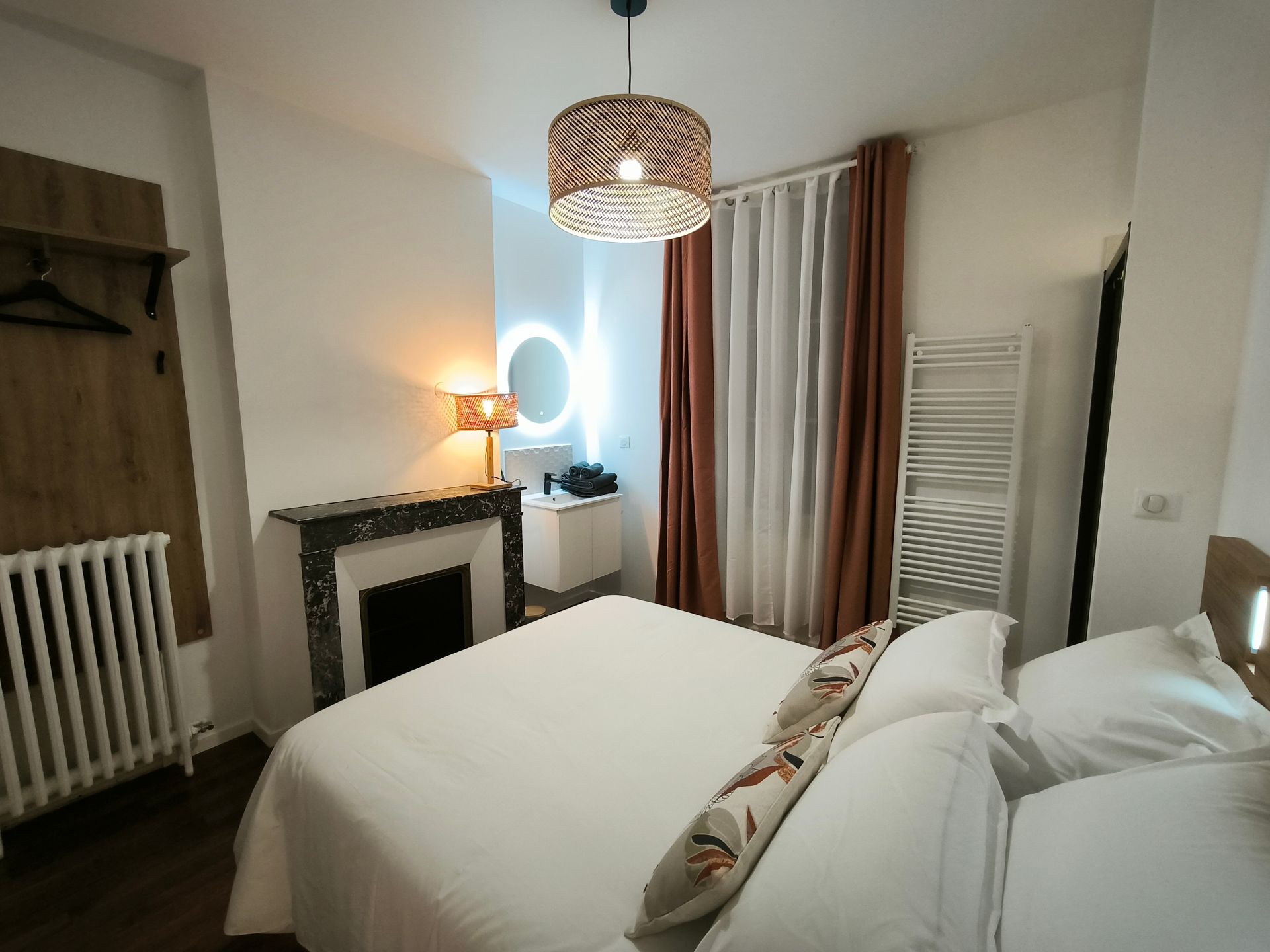 Chambre confortable avec lit blanc, cheminée, abat-jour à motifs, radiateur et fenêtre avec rideaux.