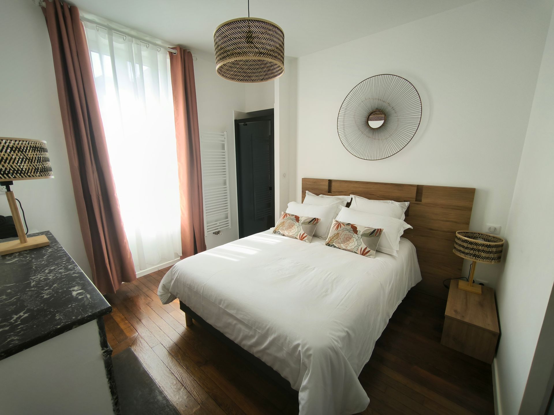Chambre lumineuse avec un lit en bois, des suspensions lumineuses en fibres naturelles, des murs blancs et la lumière du soleil filtrant à travers des voilages.