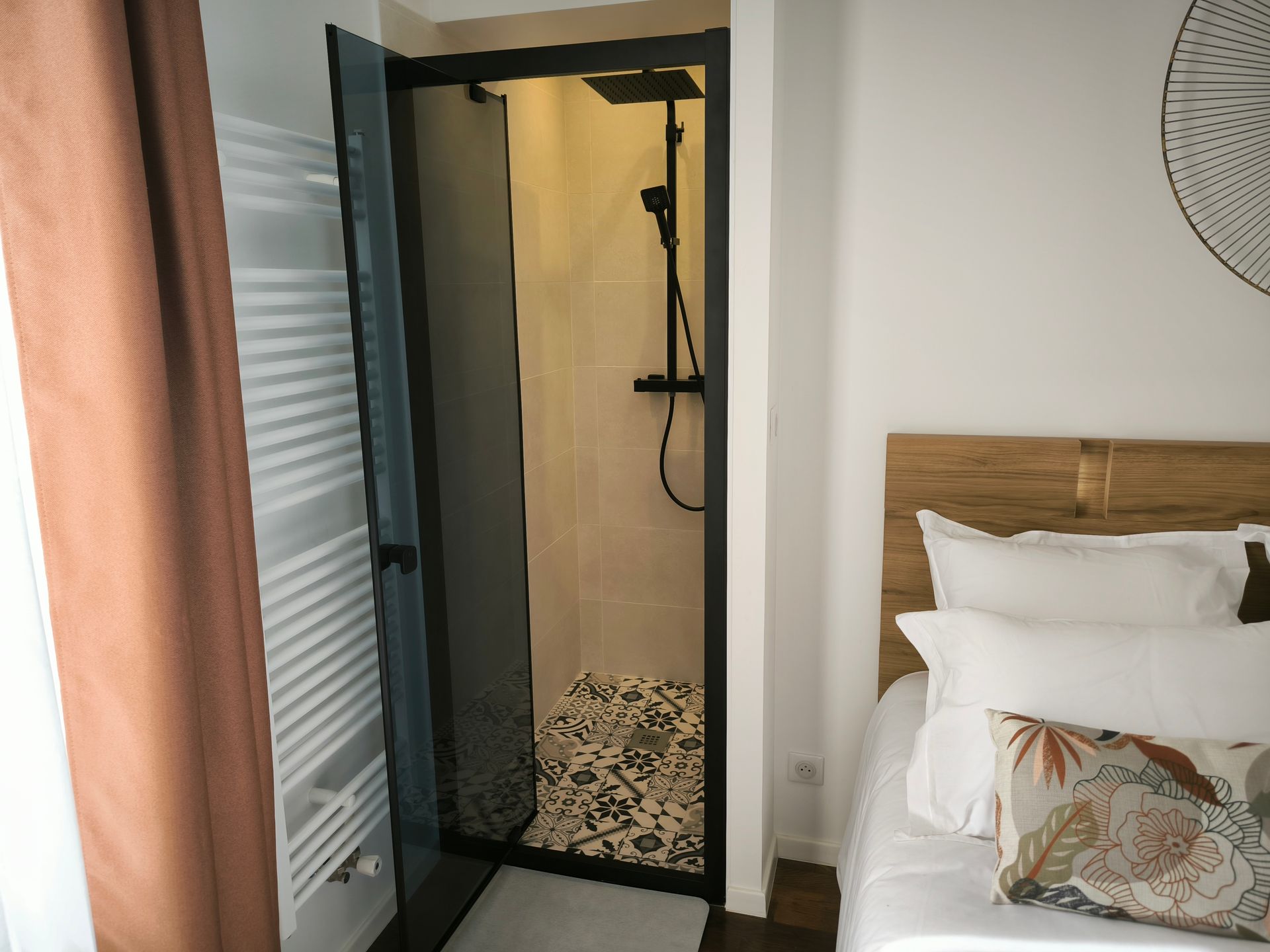 Petite chambre avec salle de douche ouverte, lit en bois et ventilateur mural en osier.