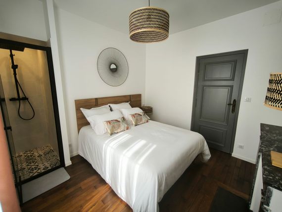 Petite chambre avec lit blanc, tête de lit en bois, porte grise, salle de douche noire et plafonnier en tissu.