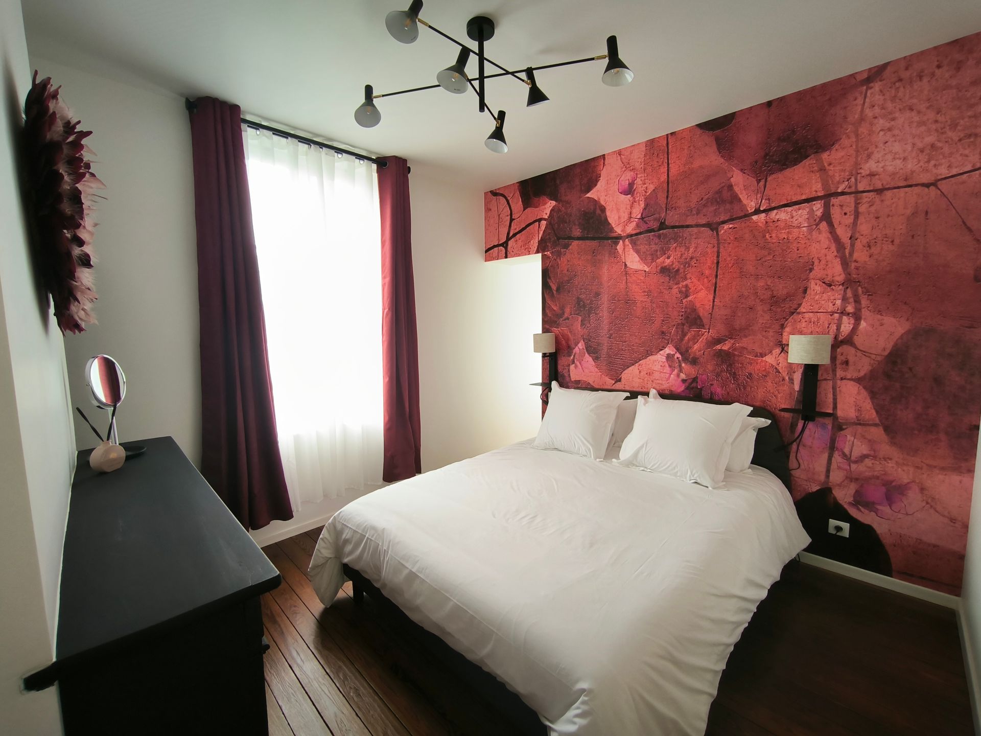 Chambre avec un mur d'accent rose, un lit blanc, des rideaux foncés et une commode noire sous un plafonnier.