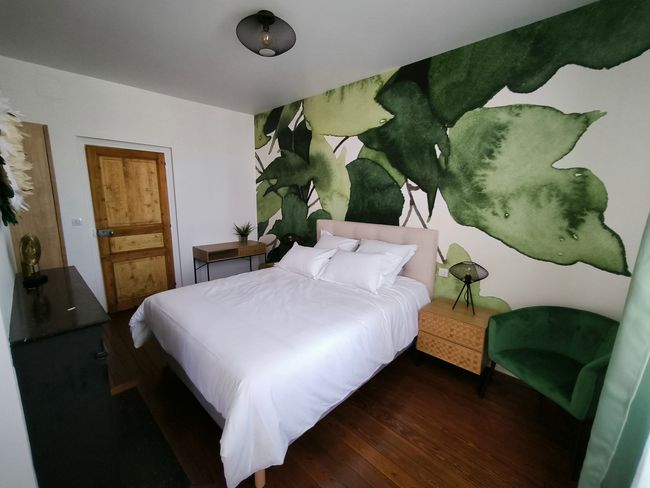 Chambre confortable avec un lit blanc, une fresque murale de feuilles vertes, un parquet et une porte en bois