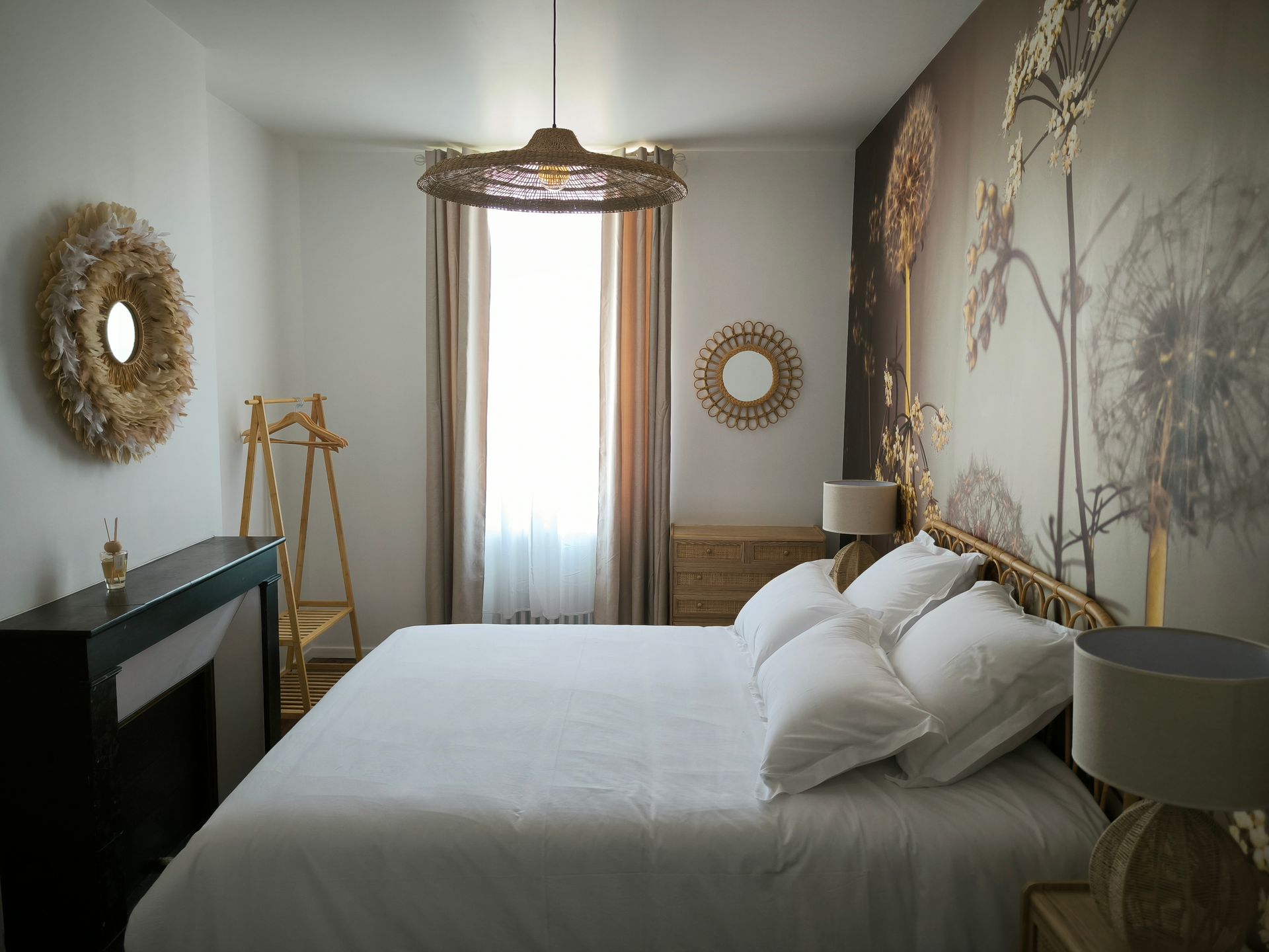Chambre lumineuse avec un lit blanc, une fresque murale à motifs et une fenêtre encadrée de rideaux et de décorations.