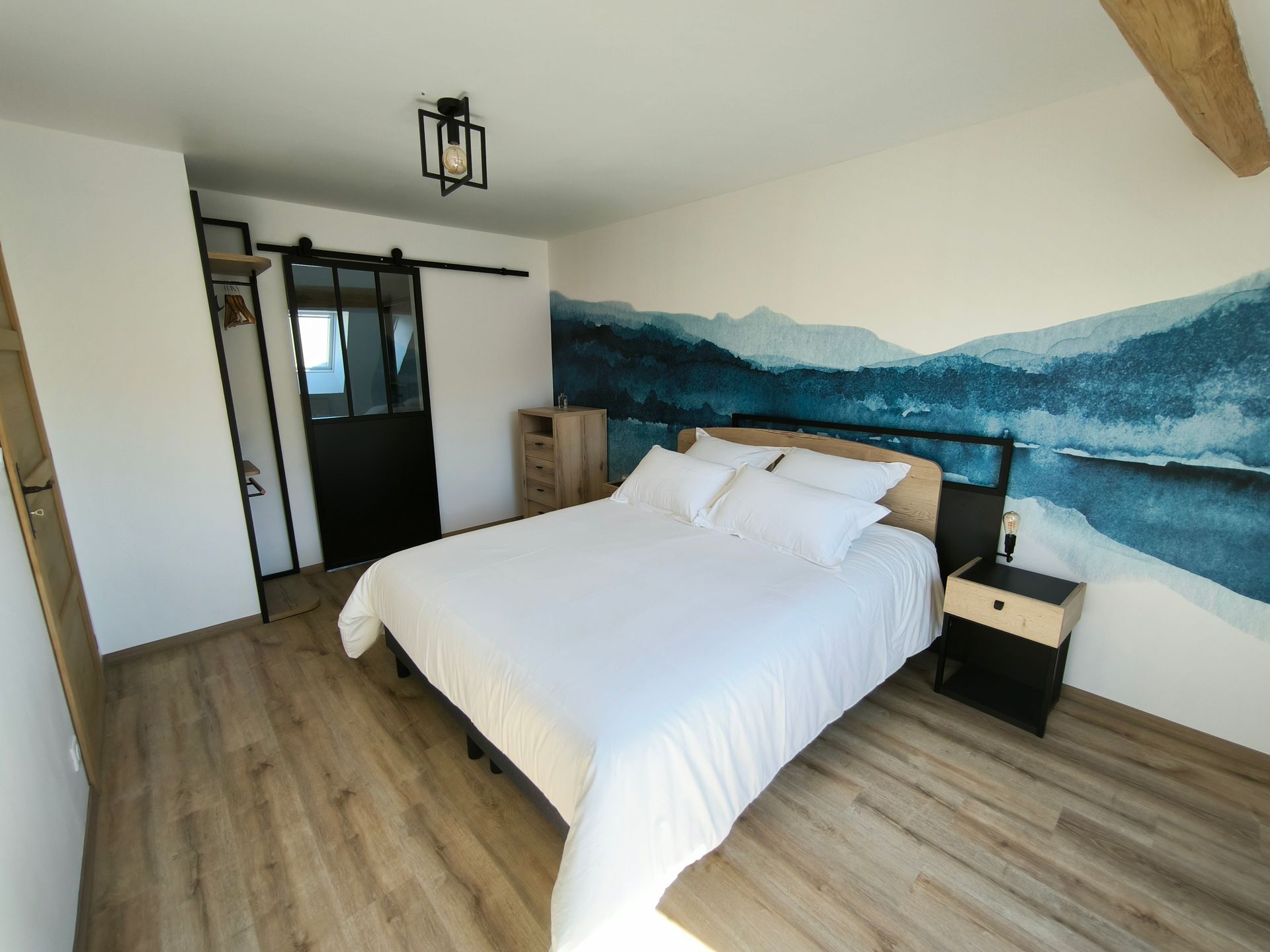 Chambre moderne avec un lit blanc, une fresque murale représentant une montagne bleue et un dressing ouvert.