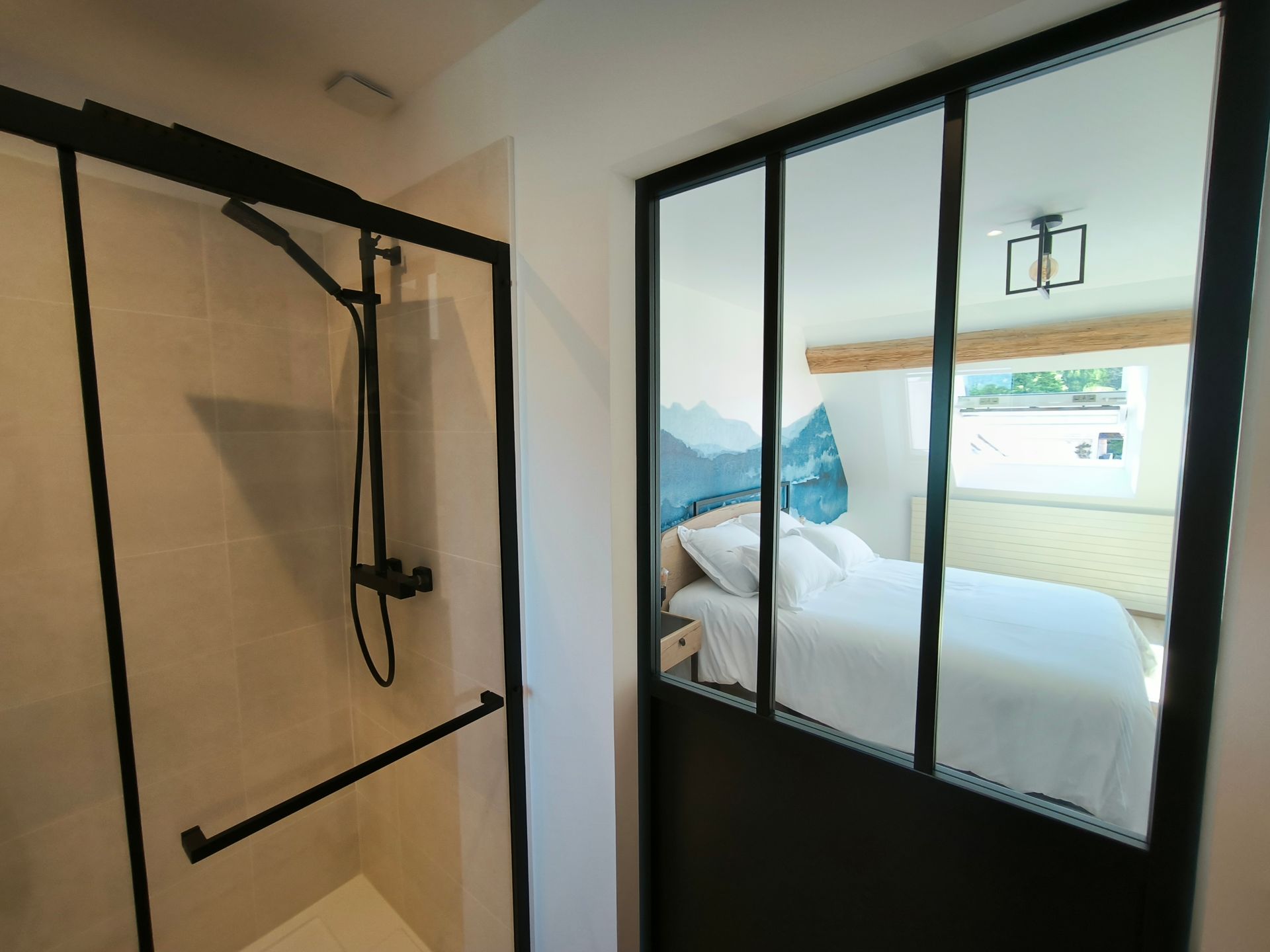 Chambre moderne avec une cloison vitrée à cadre noir et une vue sur un lit soigneusement fait près d'une fenêtre.