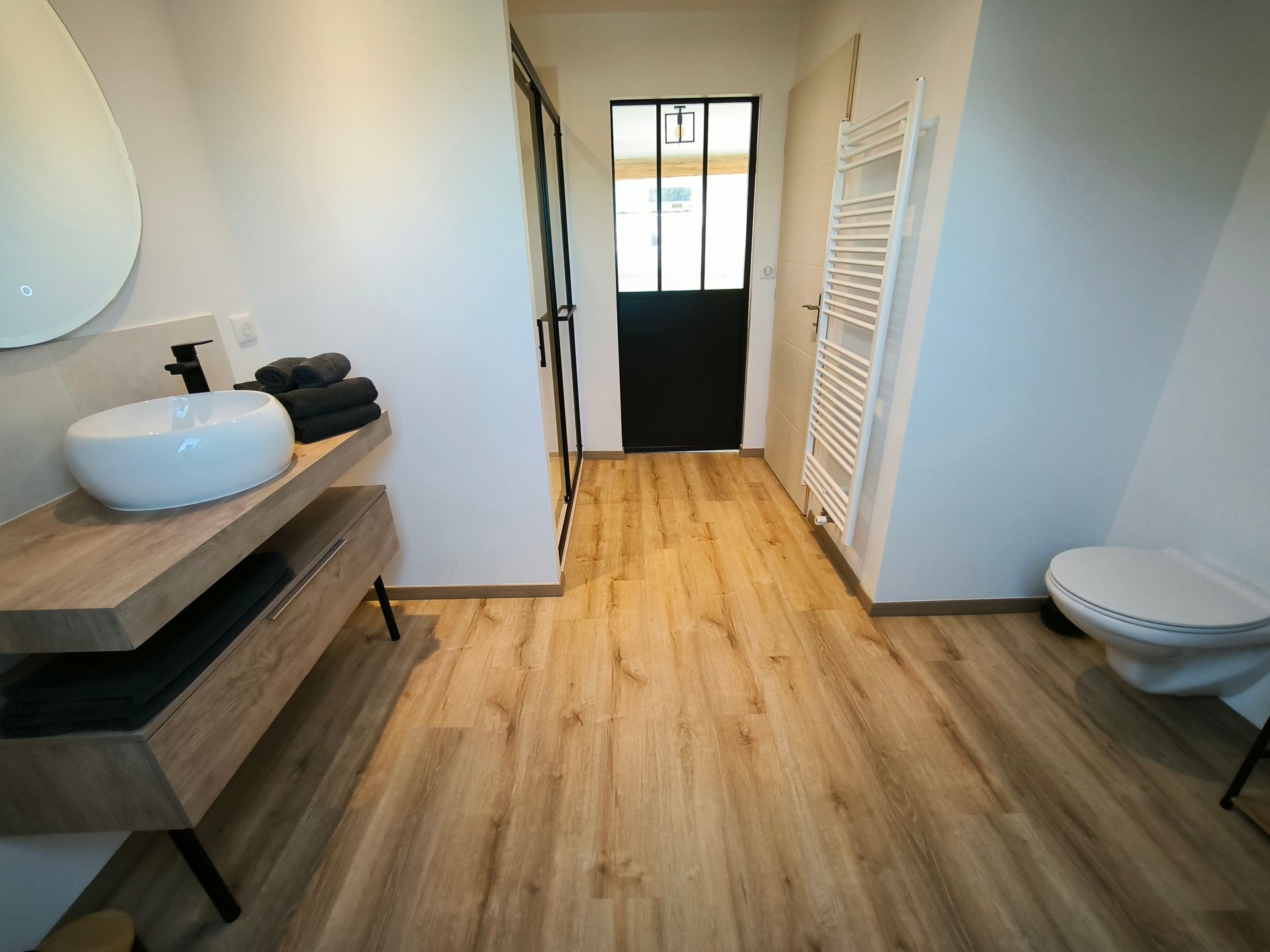 Salle de bains moderne avec meuble-lavabo en bois, lavabo rond, porte de douche à cadre noir et murs blancs.