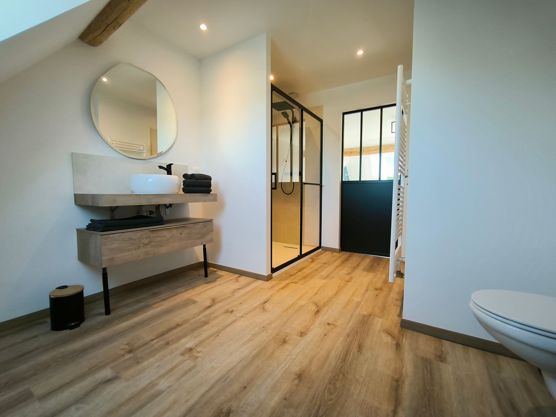 Salle de bains moderne avec parquet, meuble-lavabo mural, miroir rond et porte de douche en verre à cadre noir