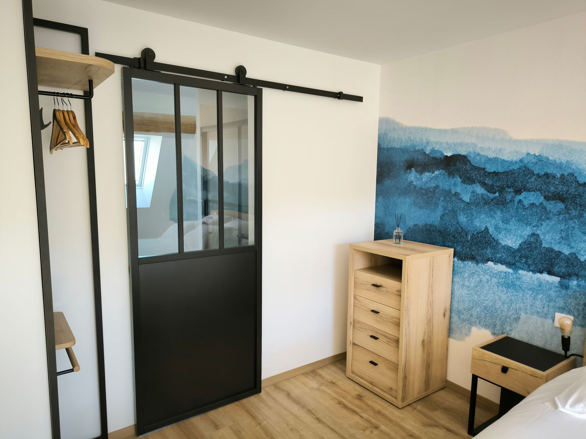 Petite chambre avec porte coulissante noire, commode en bois, bureau et fresque murale représentant une montagne bleue.