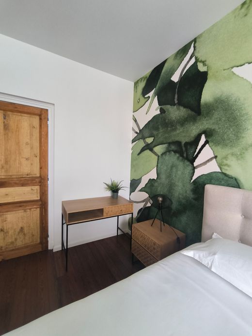 Chambre avec une grande fresque murale de feuilles vertes, une porte en bois, un bureau et un lit avec des draps blancs.