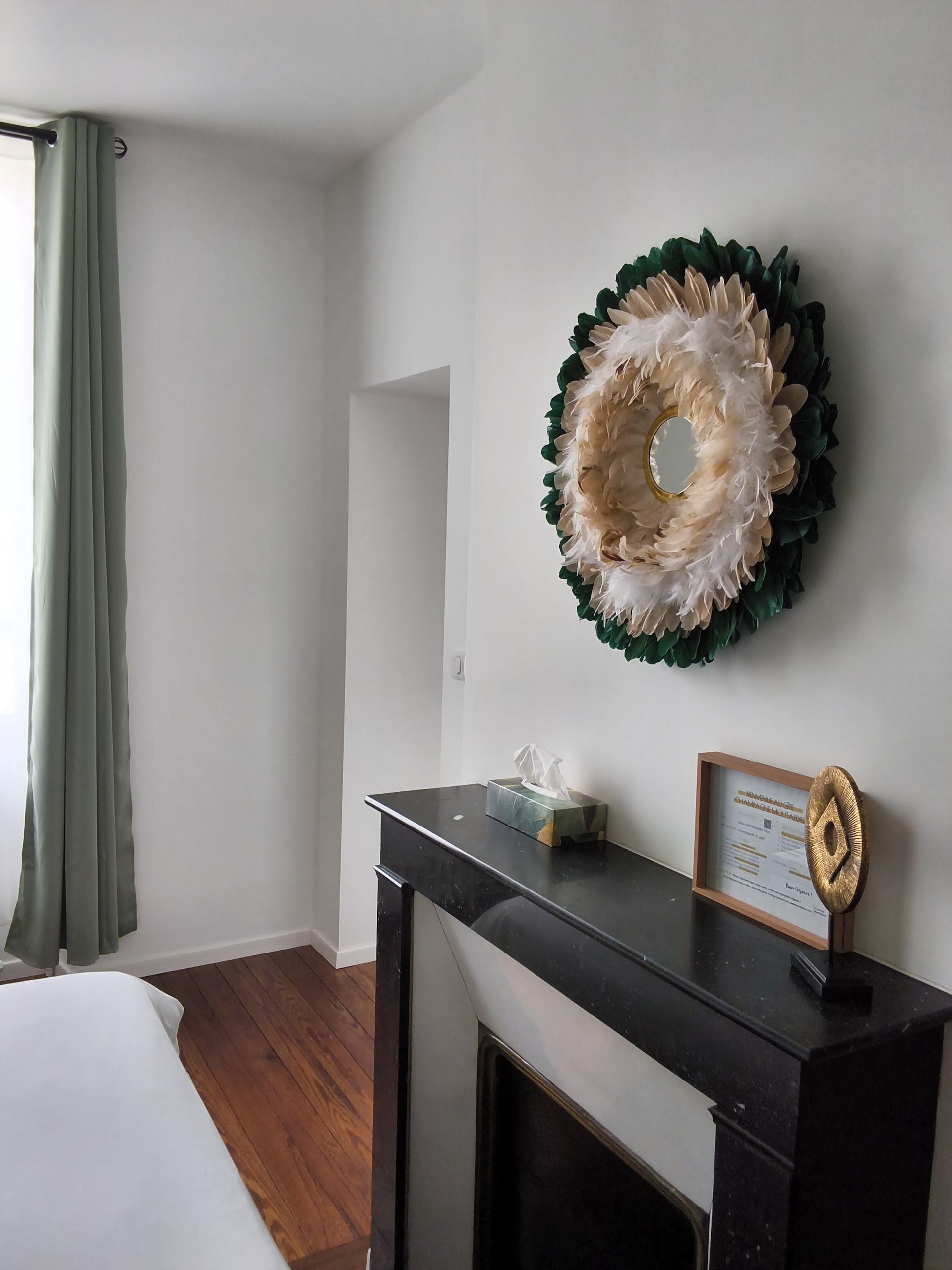 Chambre minimaliste avec une cheminée noire, un miroir décoratif et des rideaux verts au pied du lit.