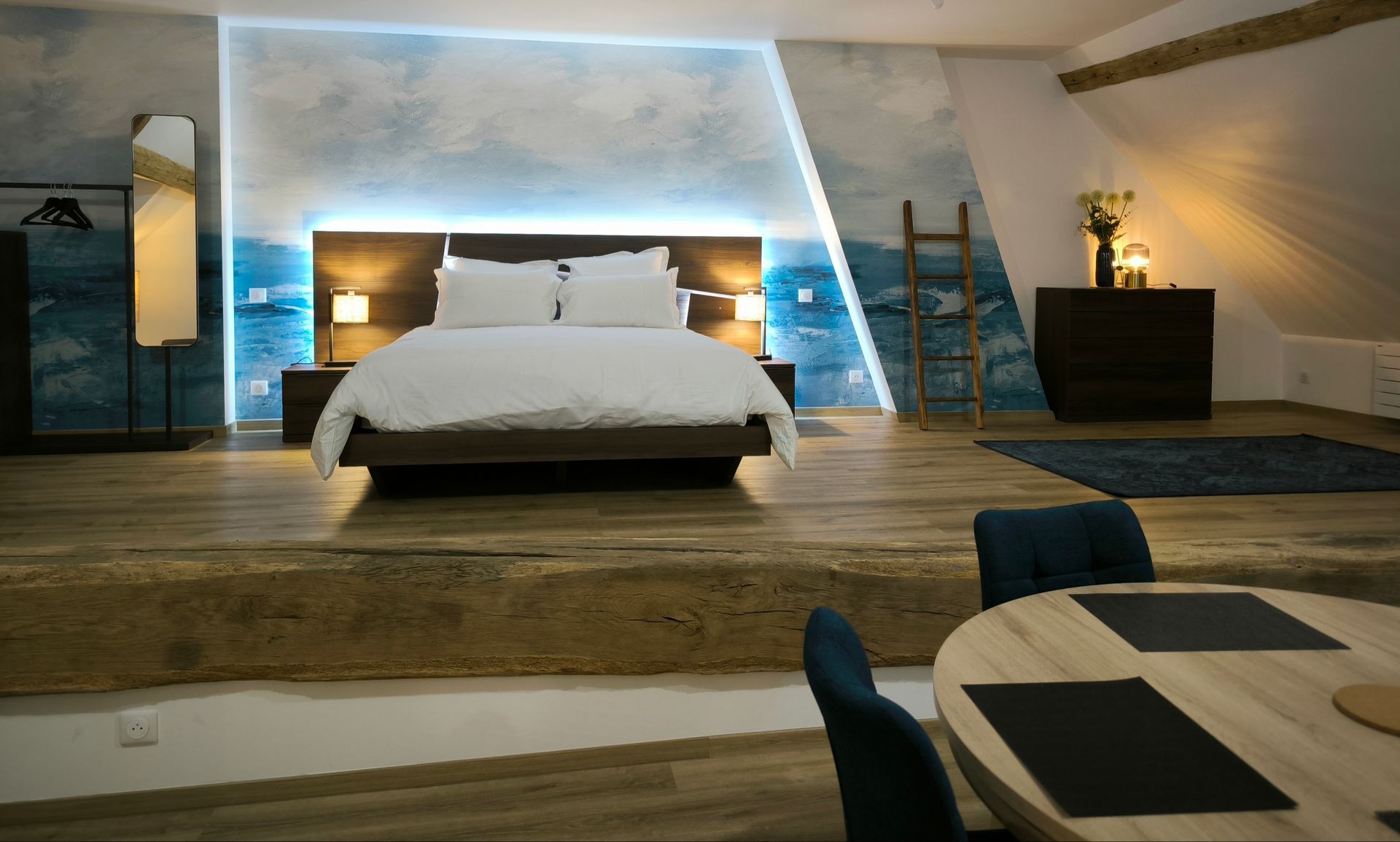 Chambre loft moderne avec lit blanc, éclairage d'ambiance bleu, estrade en bois et échelle menant à une mezzanine.