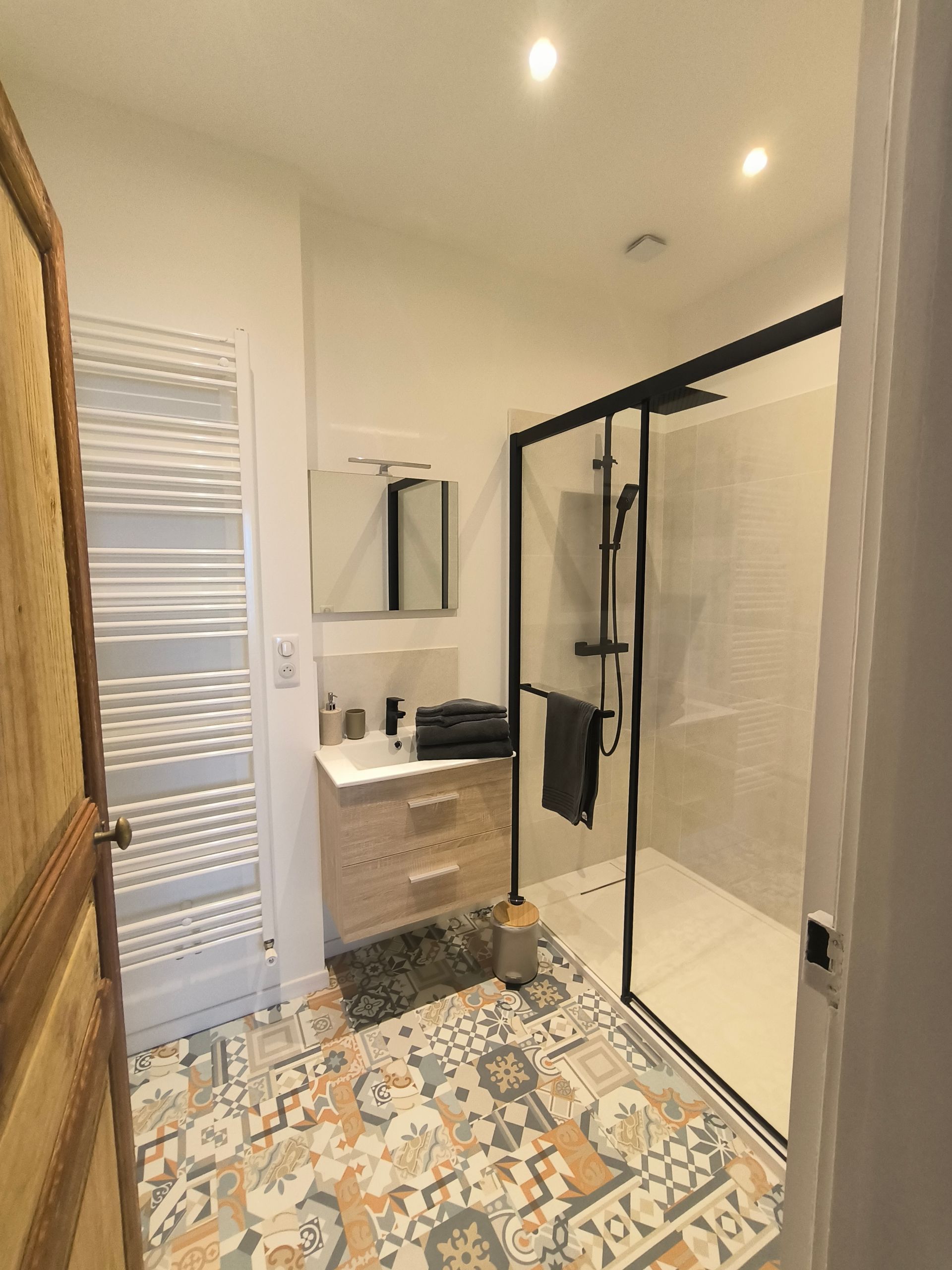 Petite salle de bains avec carrelage au sol à motifs, meuble-lavabo, miroir, sèche-serviettes et cabine de douche en verre.