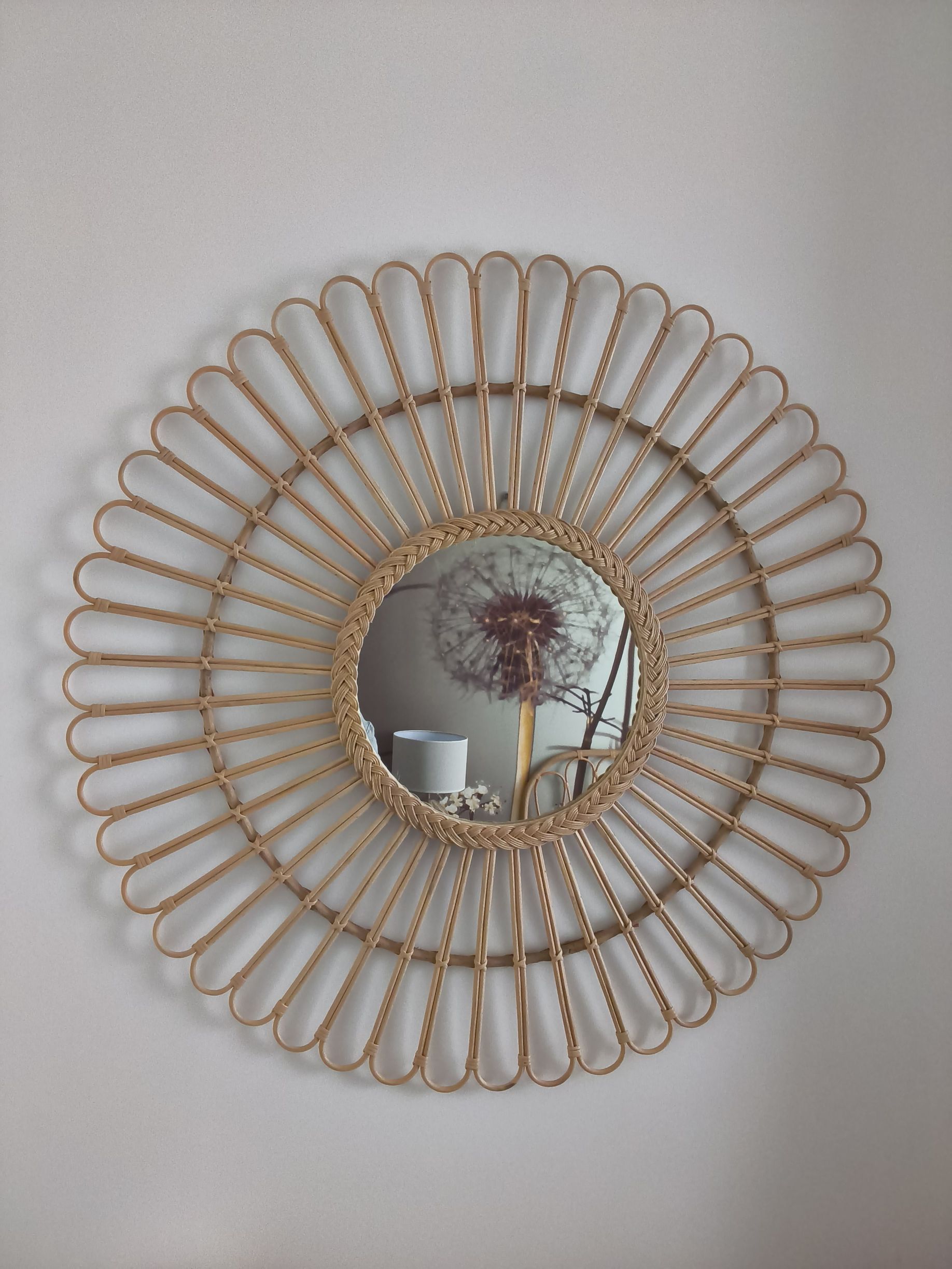 Miroir rond en osier en forme de soleil, fixé sur un mur clair, reflétant un vase contenant des fleurs séchées et des bougies.