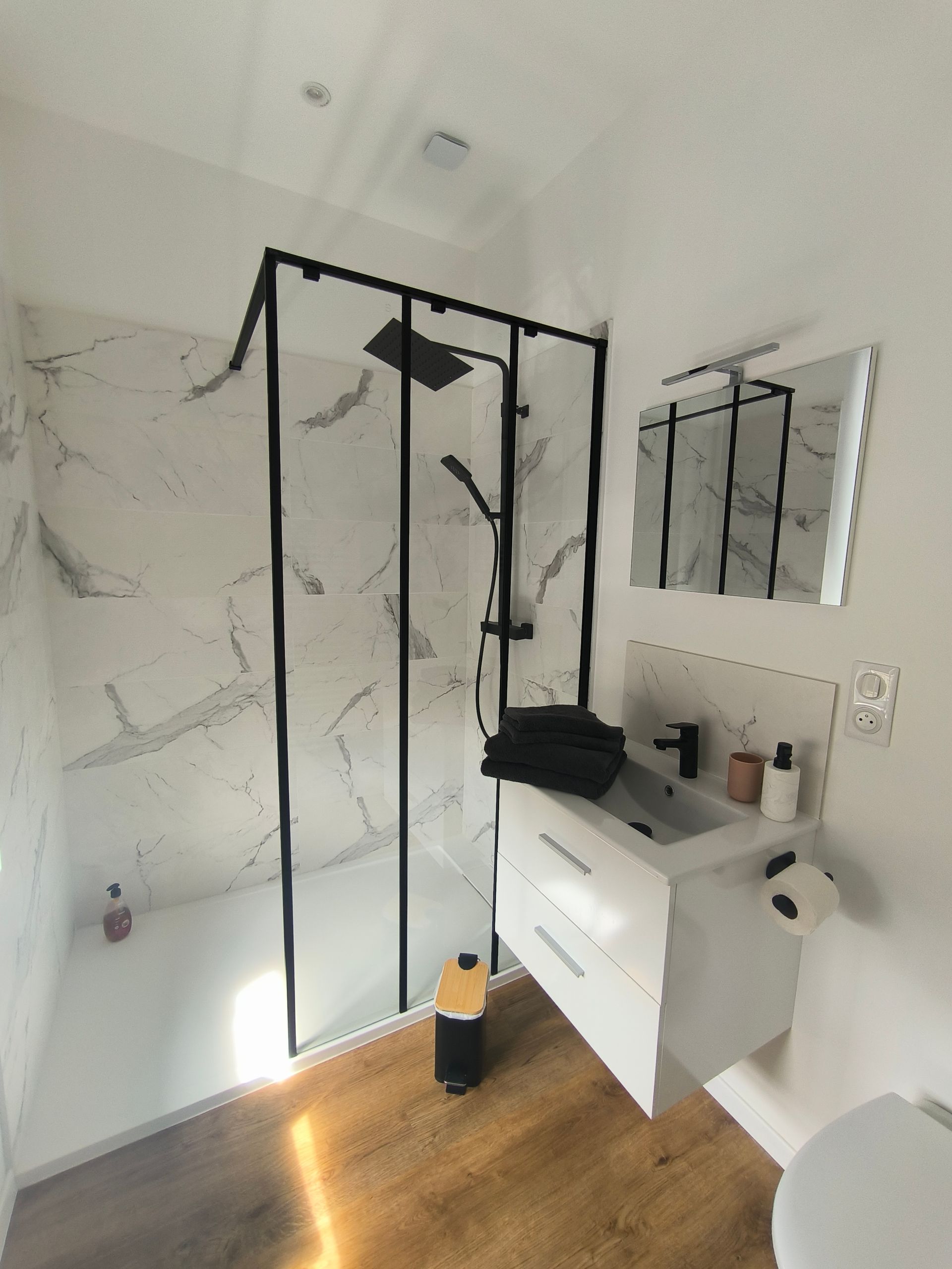 Petite salle de bains avec douche en verre à cadre noir, meuble-lavabo blanc, miroir et lumière du soleil sur le sol.