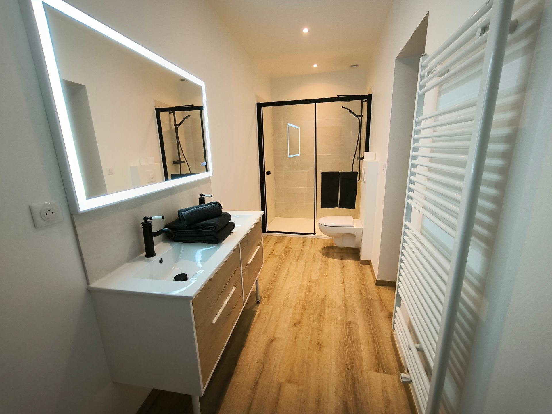 Salle de bains moderne avec meuble-lavabo, miroir éclairé, douche, toilettes et radiateur blanc sur parquet.
