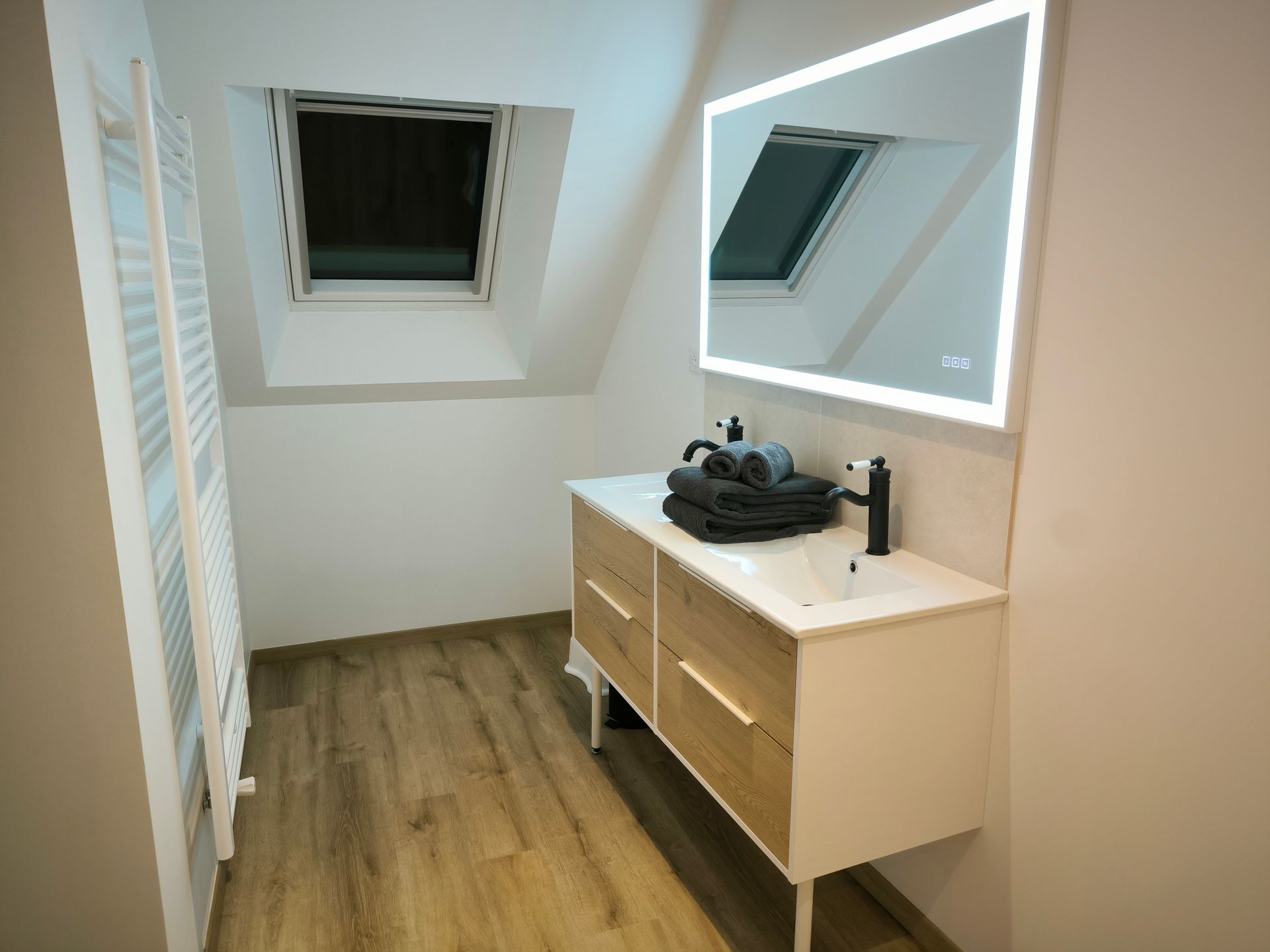 Petite salle de bain mansardée avec lavabo, miroir et armoire en bois sous fenêtres de toit.