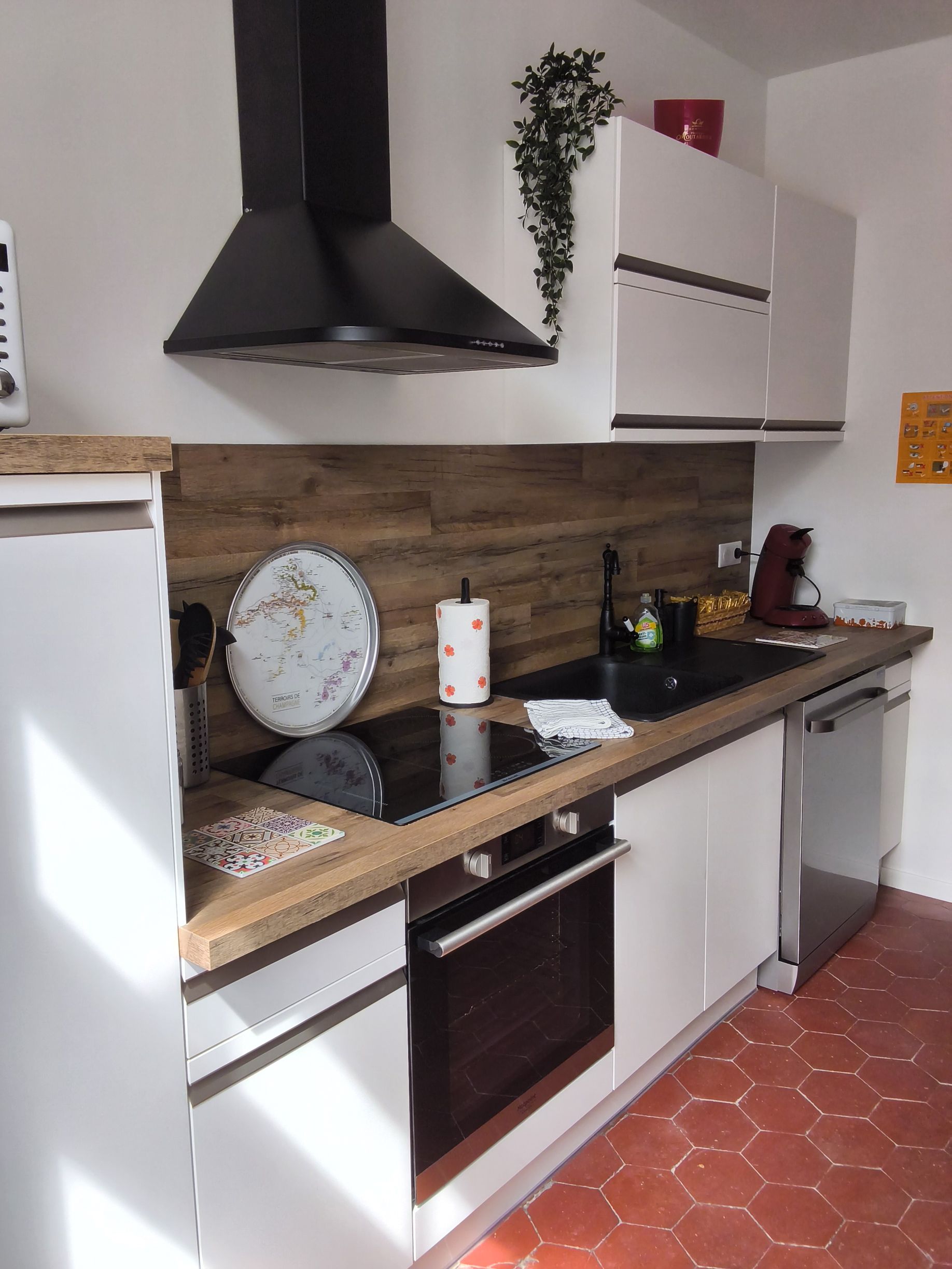 Petite cuisine moderne avec placards blancs, hotte noire, crédence en bois et sol en carrelage de terre cuite.