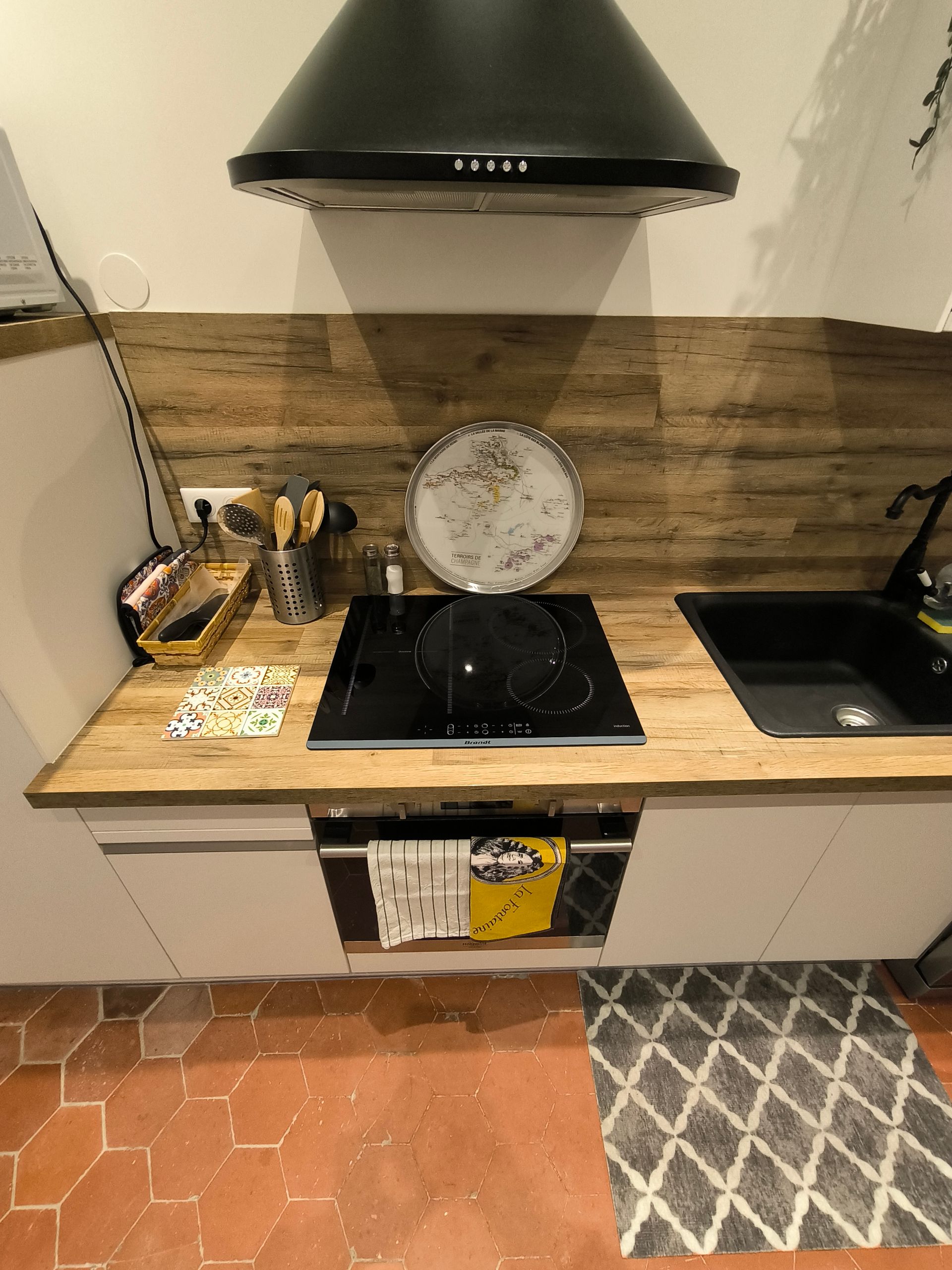 Petit comptoir de cuisine avec plaque de cuisson, hotte aspirante, évier, ustensiles et horloge, le tout adossé à une crédence en bois.