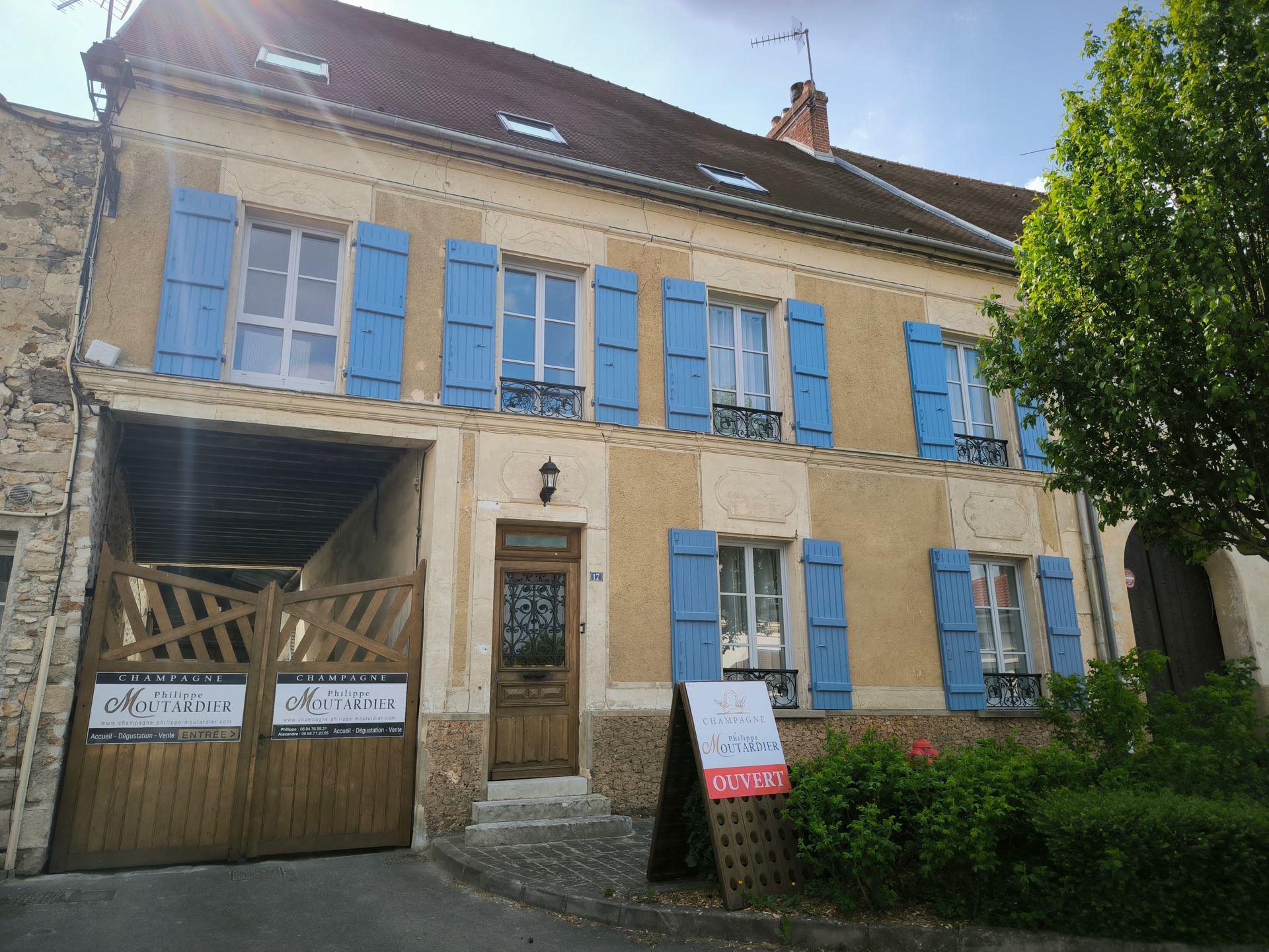 Bâtiment en pierre avec des volets bleus et une porte en bois, partiellement recouvert d'échafaudages, à côté d'un arbre feuillu.