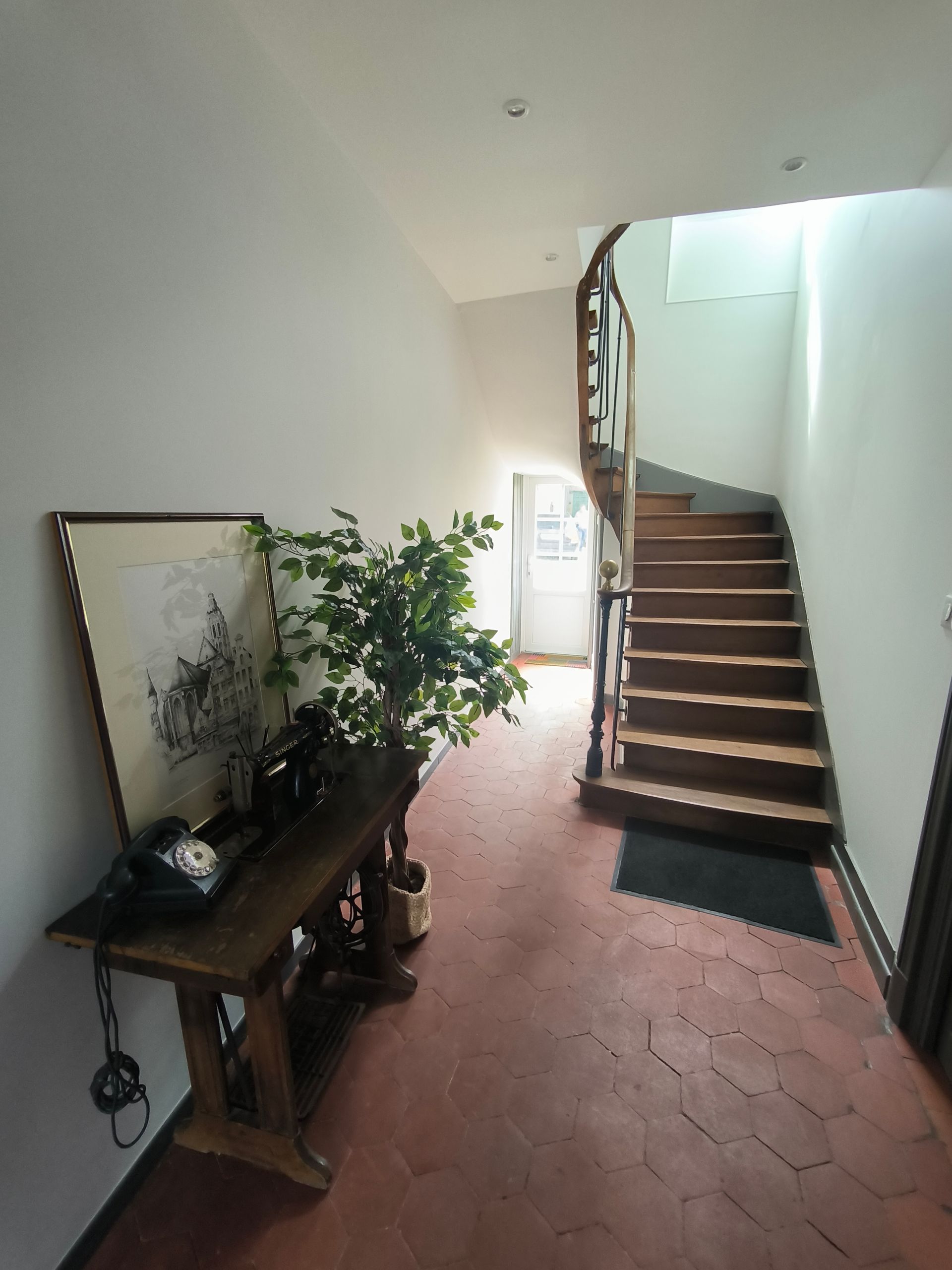 Couloir étroit avec sol en terre cuite, console en bois, miroir, plante en pot et escalier menant à l'étage.