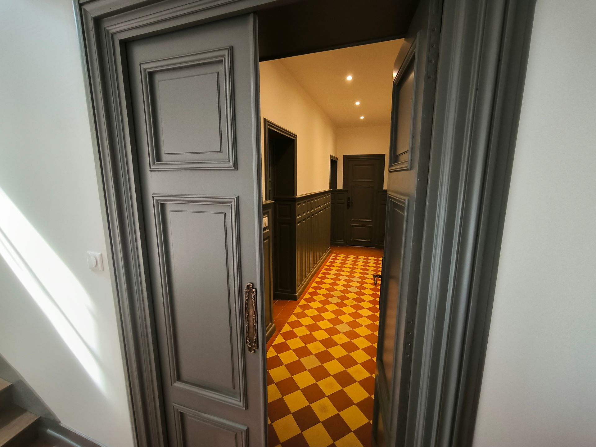 Porte ouverte donnant sur un couloir étroit avec un sol à damier jaune et rouge et des spots encastrés au plafond