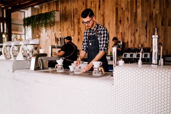 Un barista prépare du café versé sur un comptoir avec un mur en bois et du carrelage blanc.