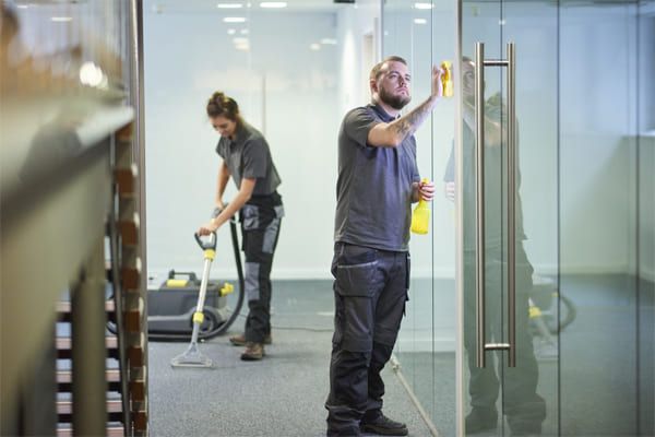 Deux personnes nettoient le bureau : l'une aspire la moquette, l'autre nettoie la porte vitrée. Intérieur lumineux.