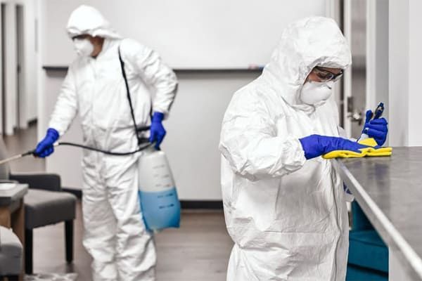 Deux personnes en combinaison blanche de protection contre les matières dangereuses désinfectent un espace intérieur. L'une vaporise, l'autre essuie une surface.