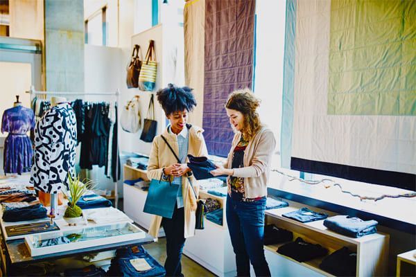 Une femme fait ses courses dans un magasin de vêtements, observant les vêtements avec une vendeuse. Pièce lumineuse avec portants et présentoirs.