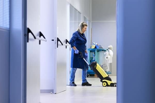 Une femme en uniforme bleu passe l'aspirateur dans un couloir de couleur claire, un chariot de nettoyage se trouvant à proximité.