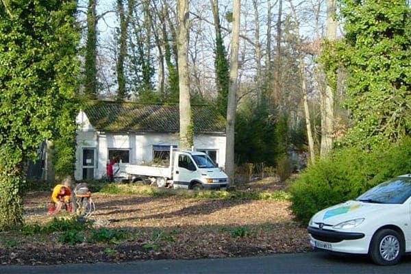 Un petit bâtiment blanc avec un camion et une voiture devant, entouré d'arbres. Des gens se tiennent près du bâtiment.