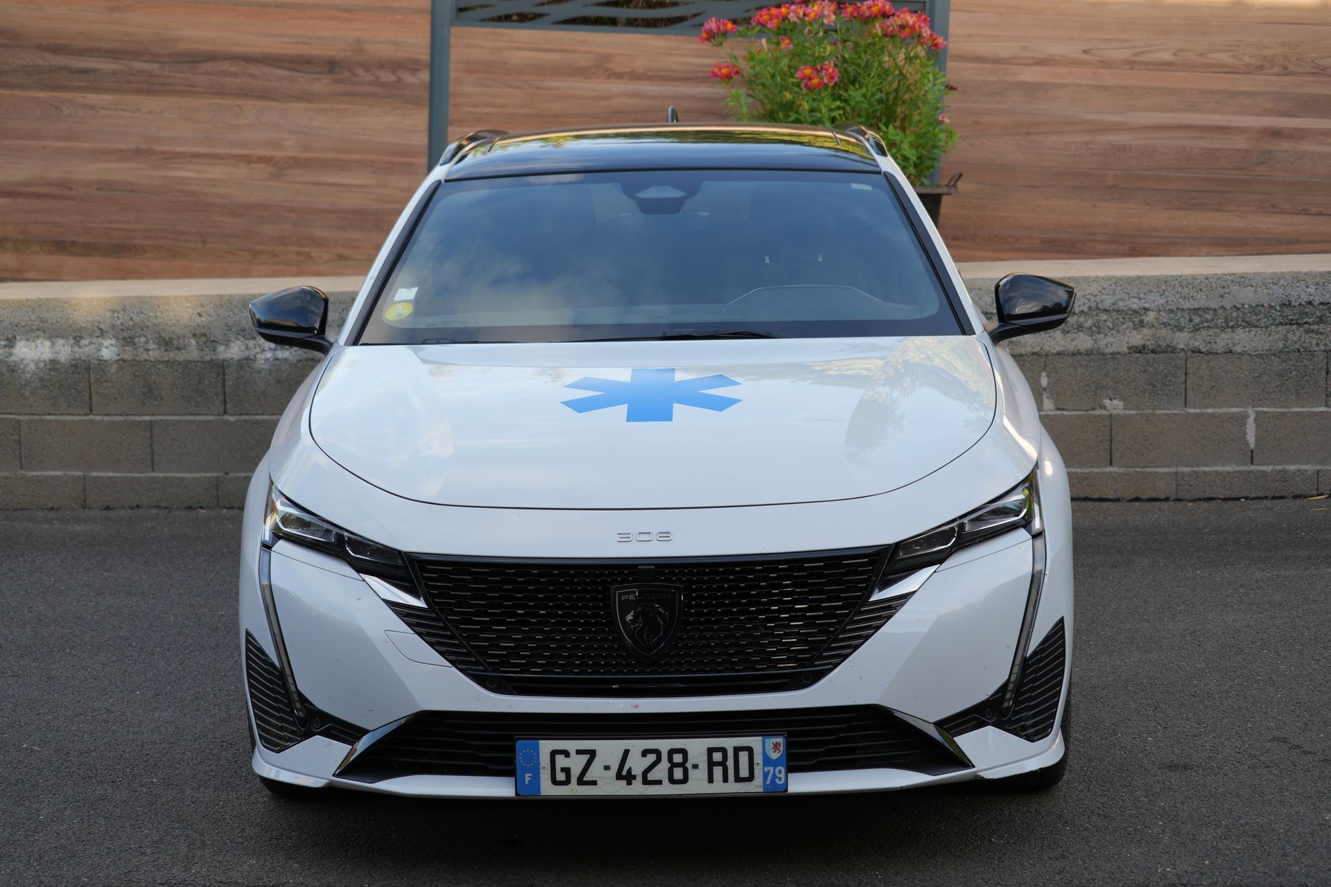 Voiture blanche d’intervention d’urgence stationnée près d’un mur au bord de la route, vue de face avec un symbole médical bleu sur le capot.