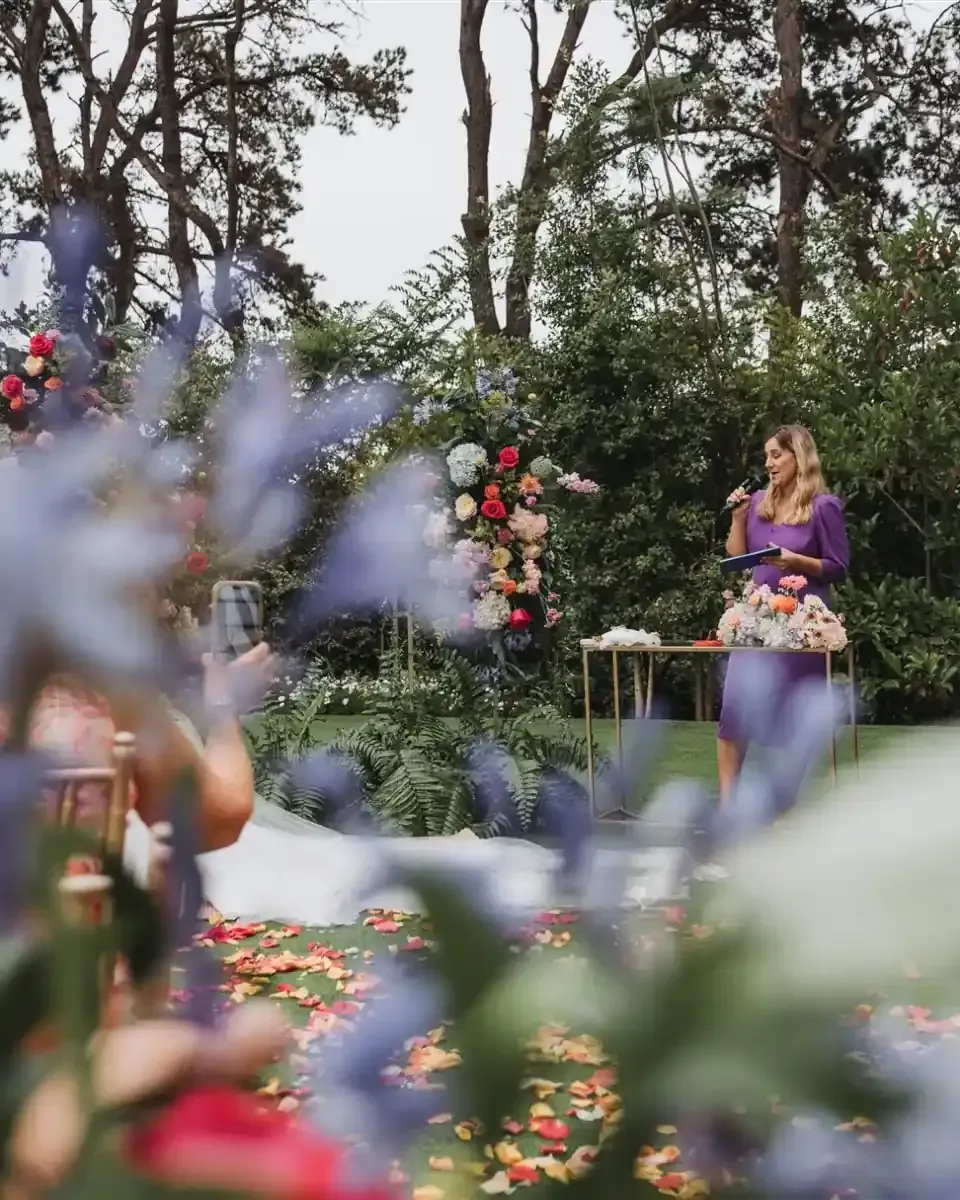 Una mujer con un vestido morado habla en una ceremonia de boda al aire libre; arco floral, fondo verde.