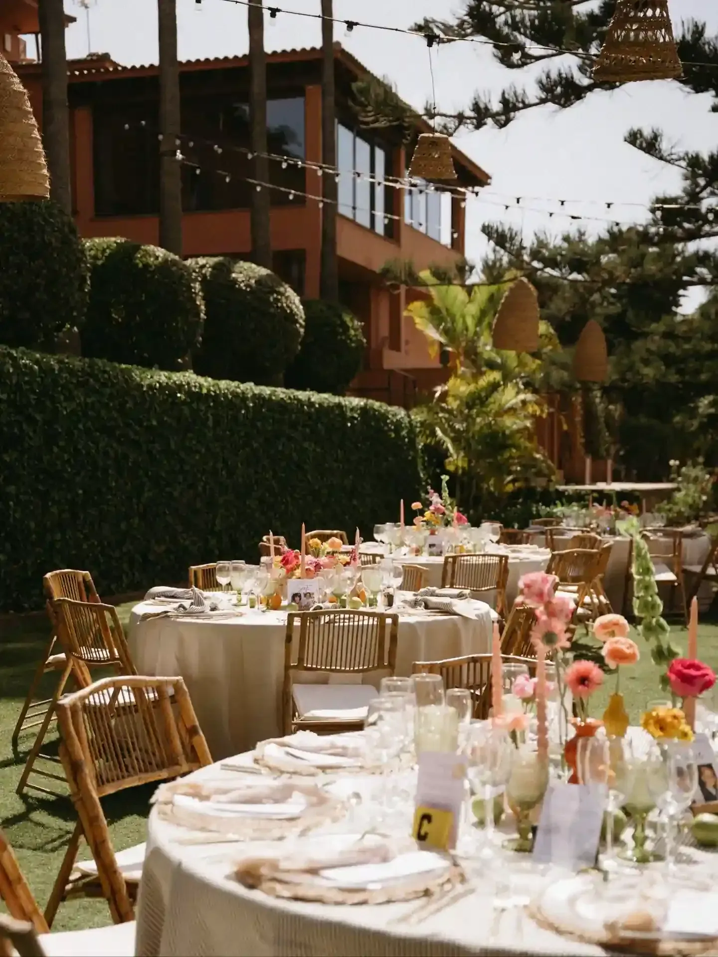 Recepción de boda al aire libre con mesas decoradas, césped verde y un edificio color terracota al fondo.