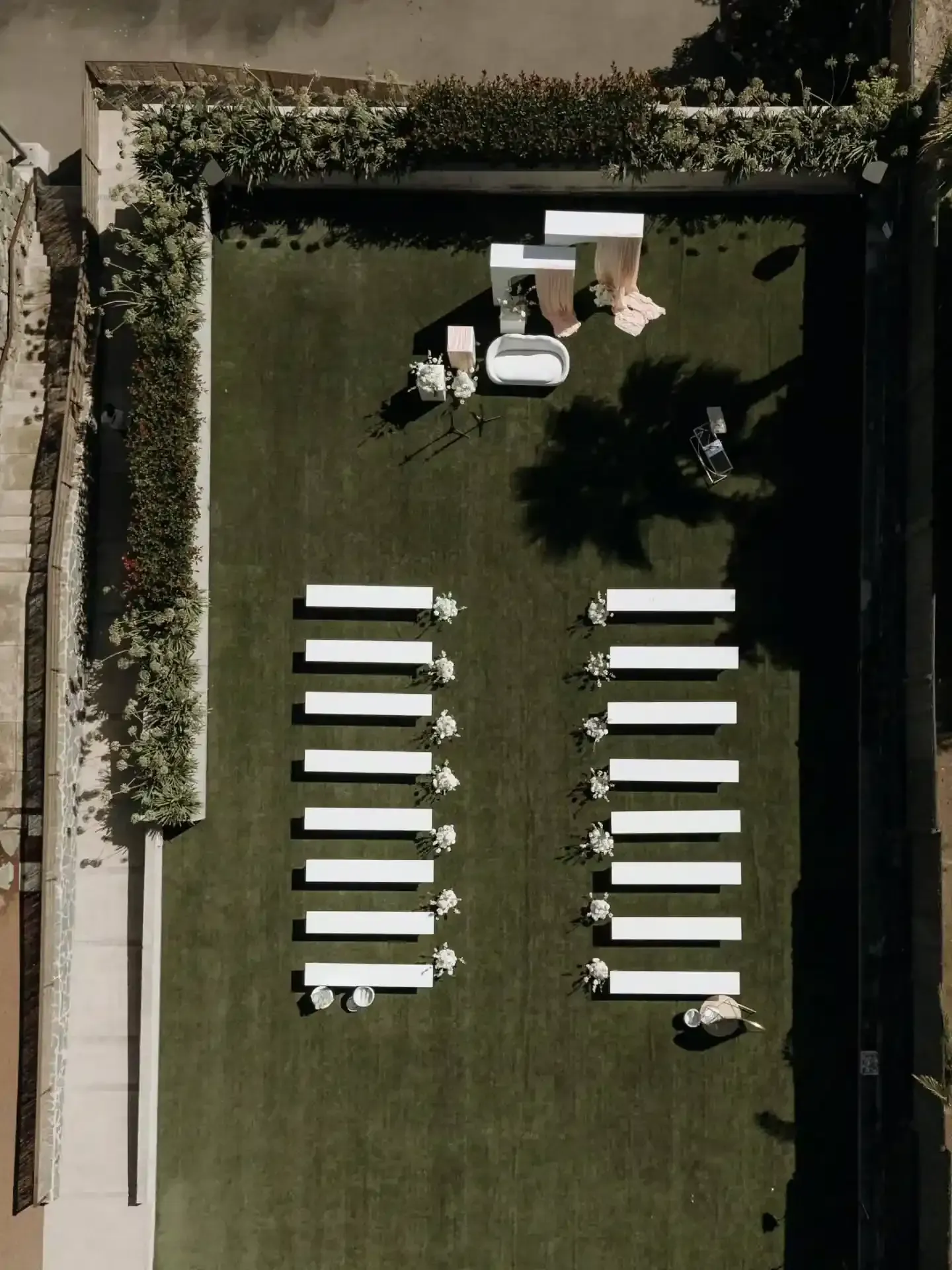 Vista aérea de un salón de bodas sobre césped verde; bancos blancos miran hacia un área de ceremonia con arreglos florales y decoraciones.