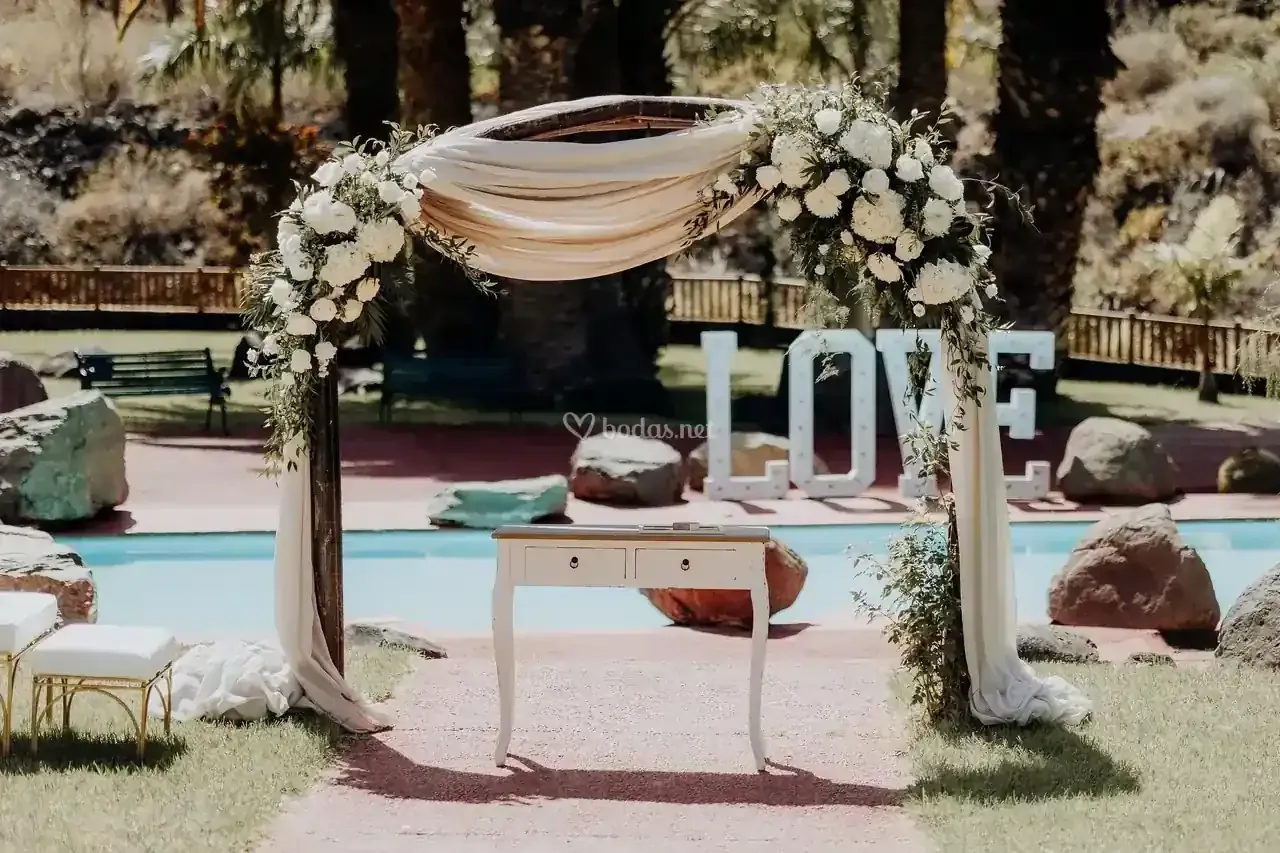 Arco de boda con flores blancas y tela drapeada, con vistas a la piscina. Letrero de