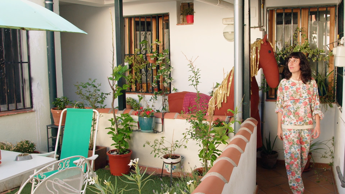 Una mujer en pijama floreado entra a un pequeño patio con jardín, con plantas en macetas, una mesa y un sillón verde.