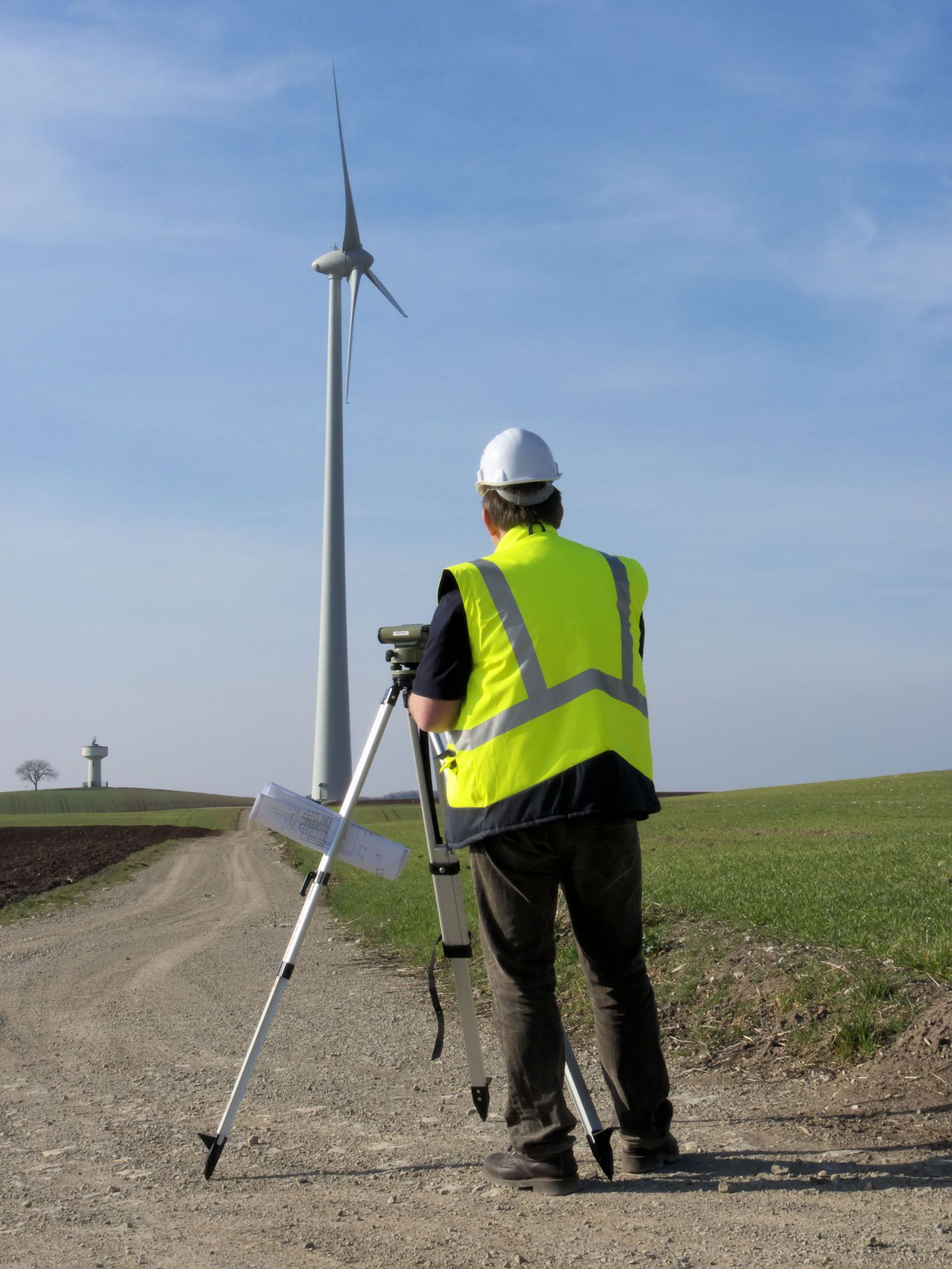 Un homme portant un gilet de sécurité inspecte une éolienne, debout sur un chemin de terre dans un champ sous un ciel bleu.