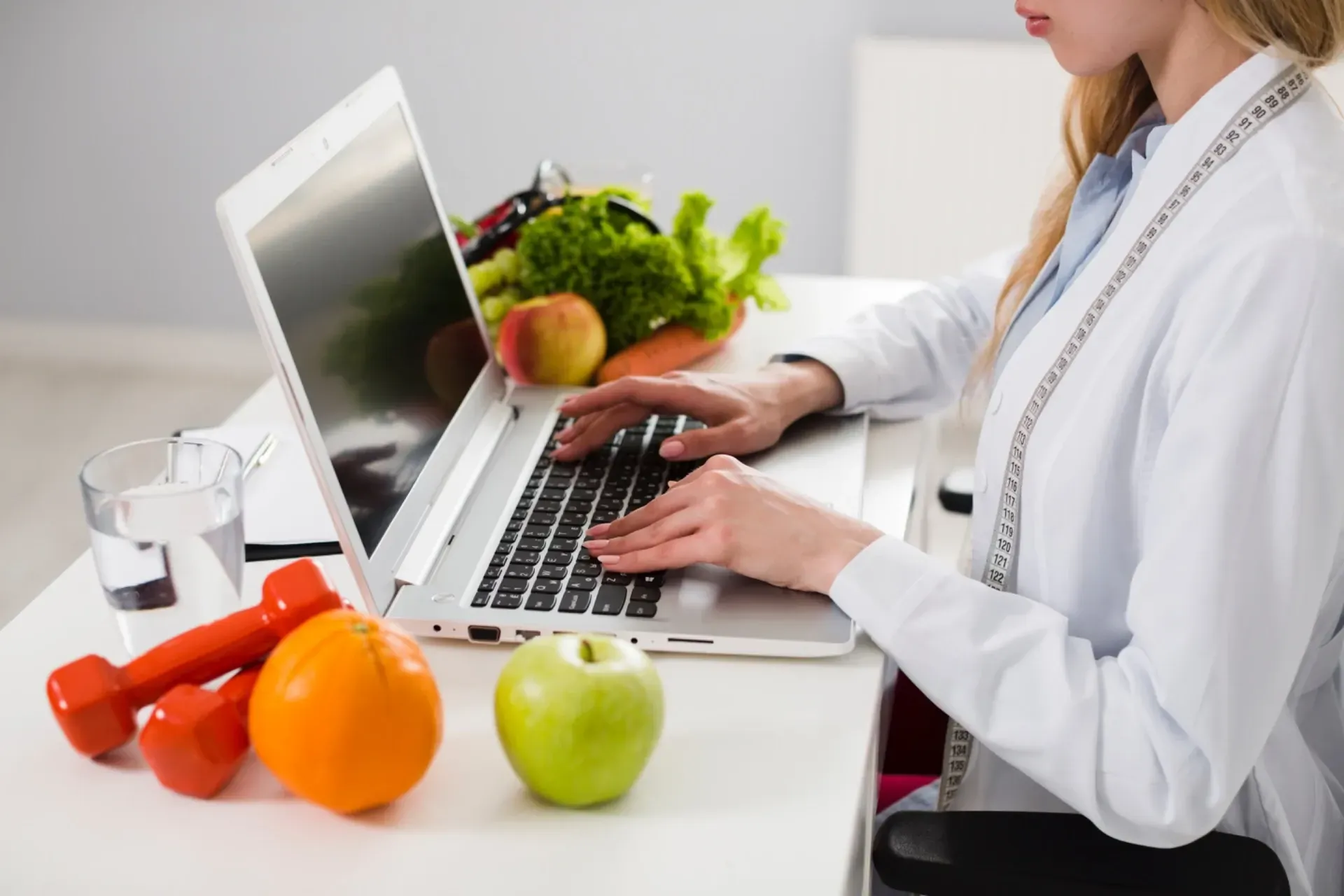 Dietista usando una computadora portátil en un escritorio con frutas, verduras, mancuernas y agua.
