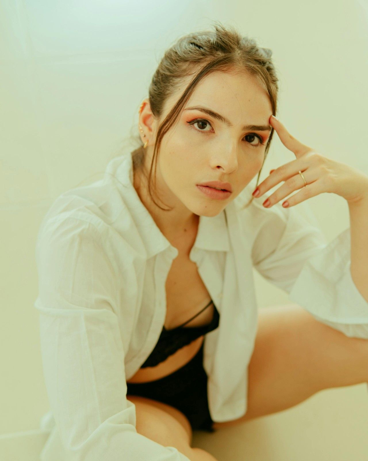 Mujer con camisa blanca y lencería negra, mano en la frente, sentada en una habitación pálida.